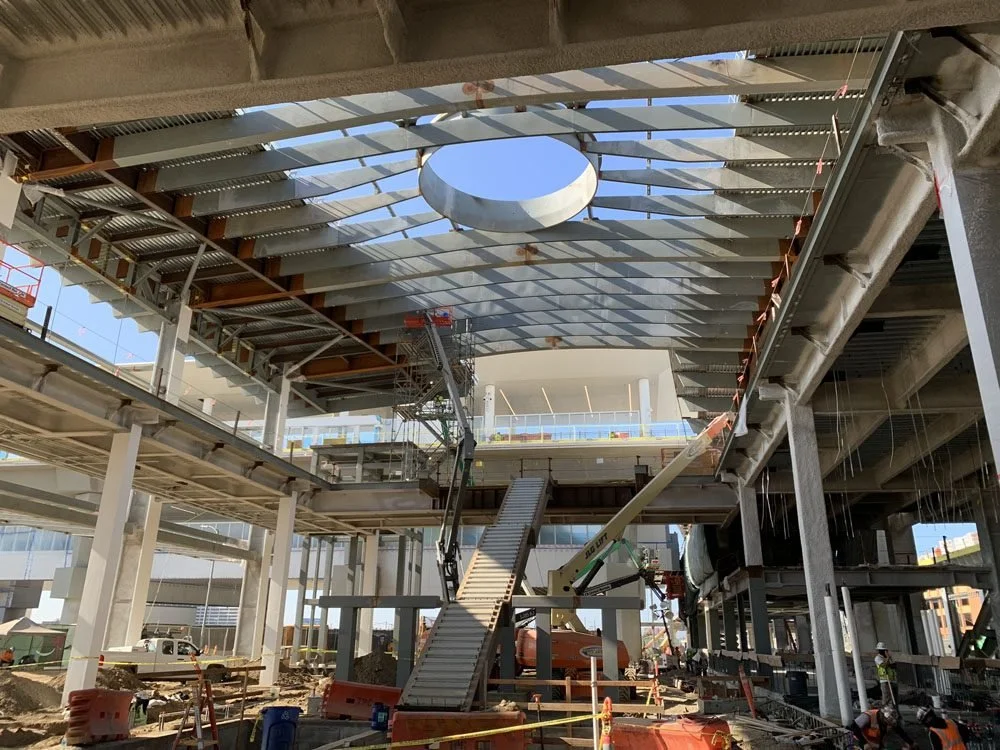 Construction site of a multi-story building with steel framework, cranes, and workers installing roofing or facade panels.