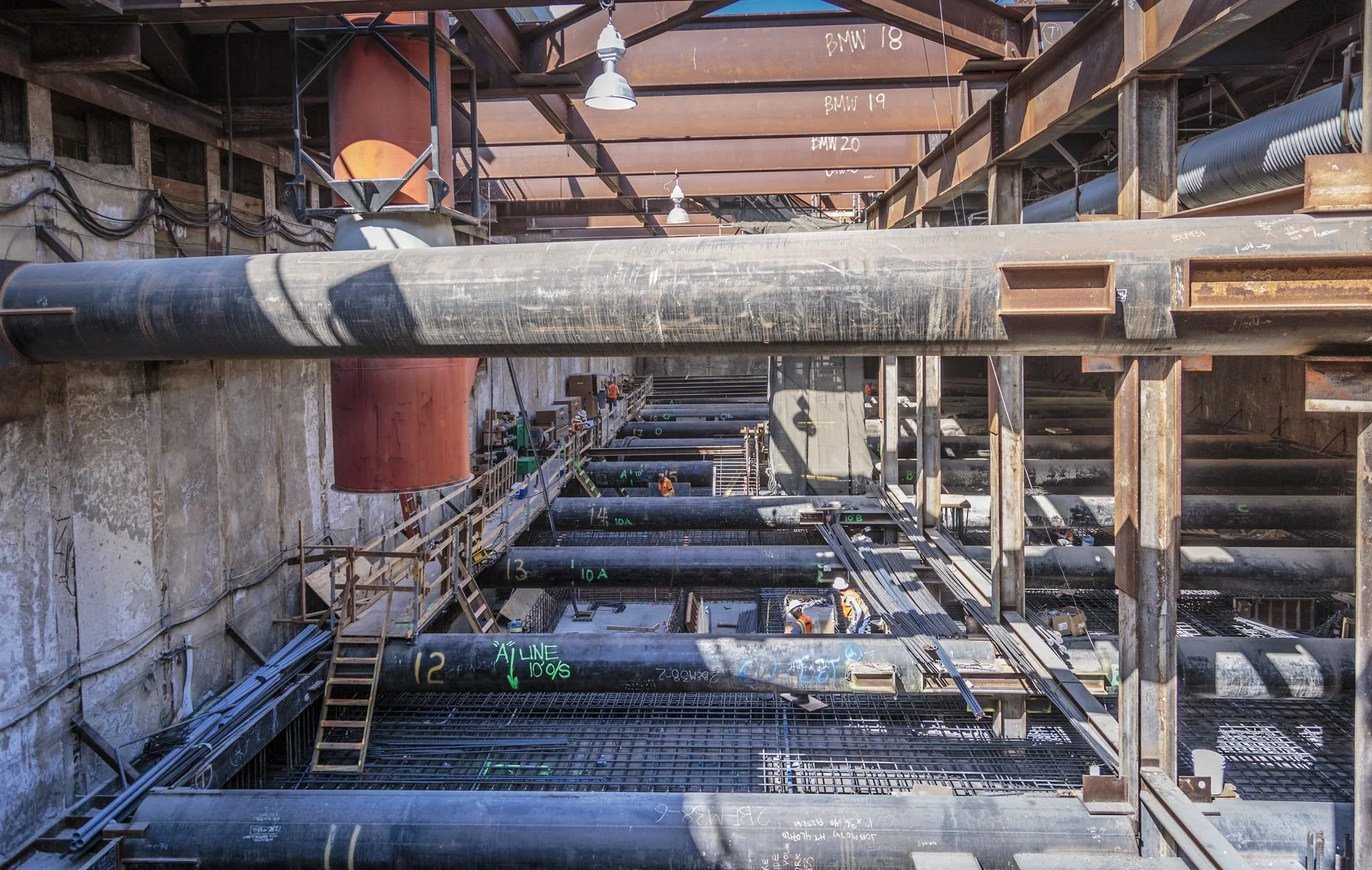 Construction site with large pipes, scaffolding, and workers in safety vests and helmets.