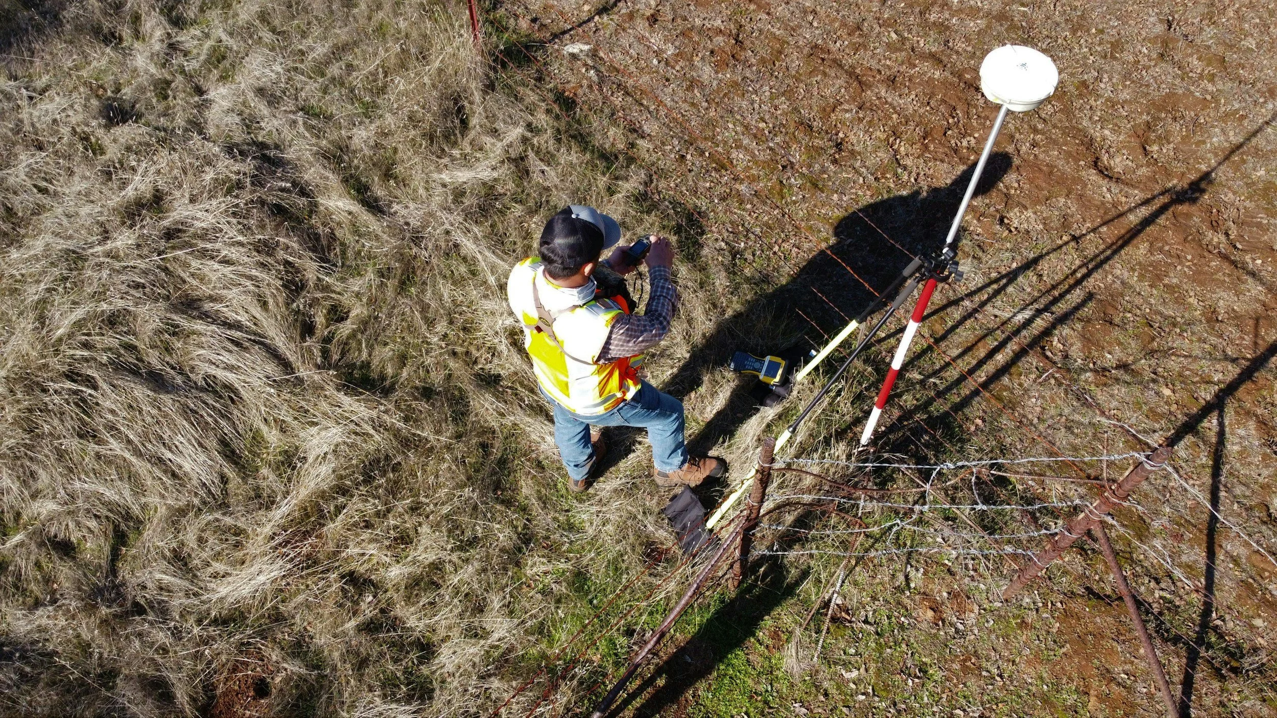 A person wearing a safety vest and hard hat working with surveying equipment on a grassy and dirt area outdoors, near a barbed wire fence.