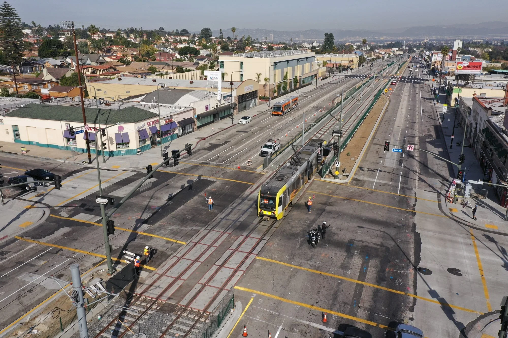 An aerial view of a city street with a light rail train, cars, construction workers, and buildings, with residential houses and hills in the background.