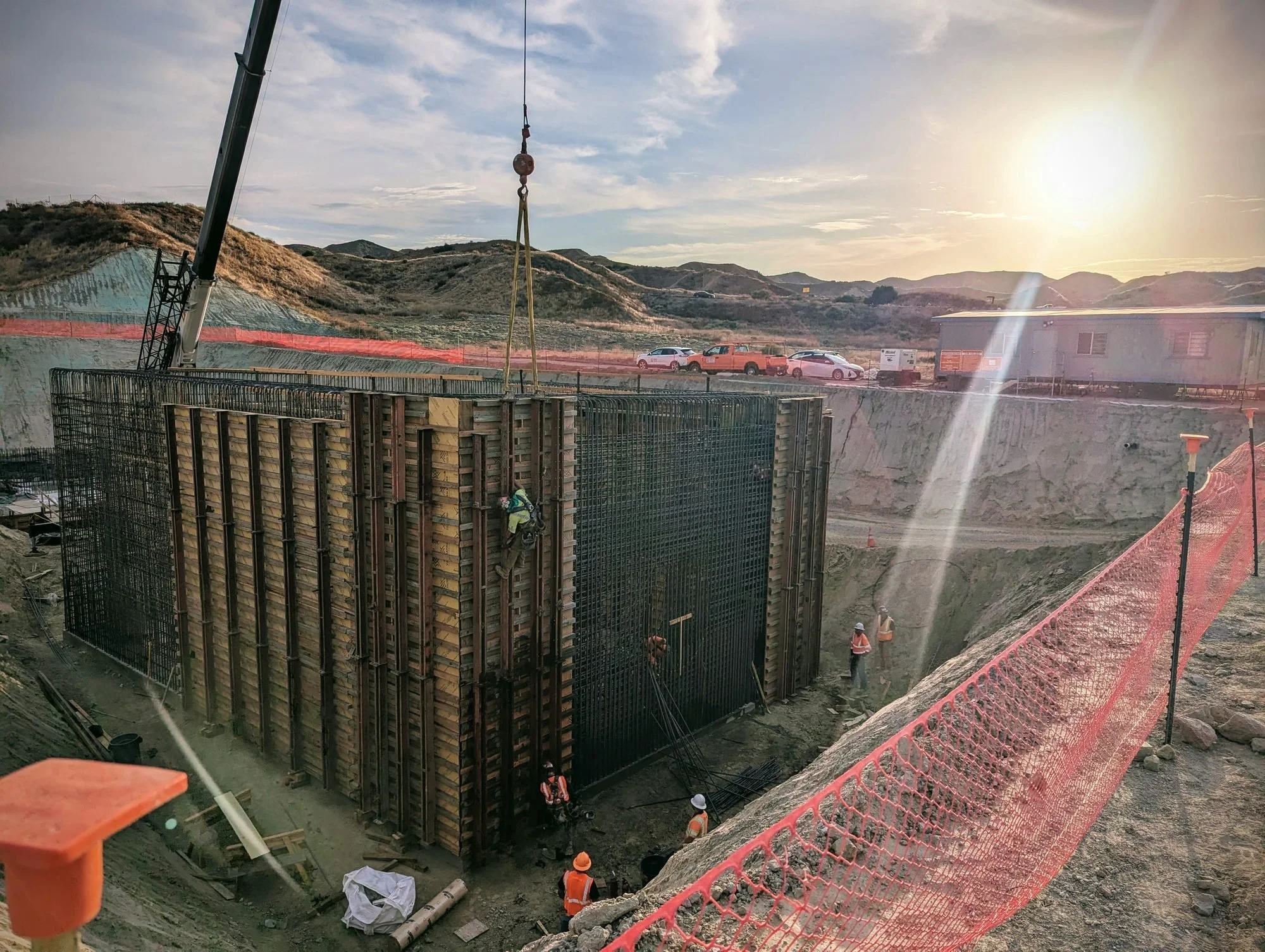 Construction workers at an active construction site, with concrete forms and reinforcement rebar, and a crane lifting materials, set against a hillside landscape during sunset.