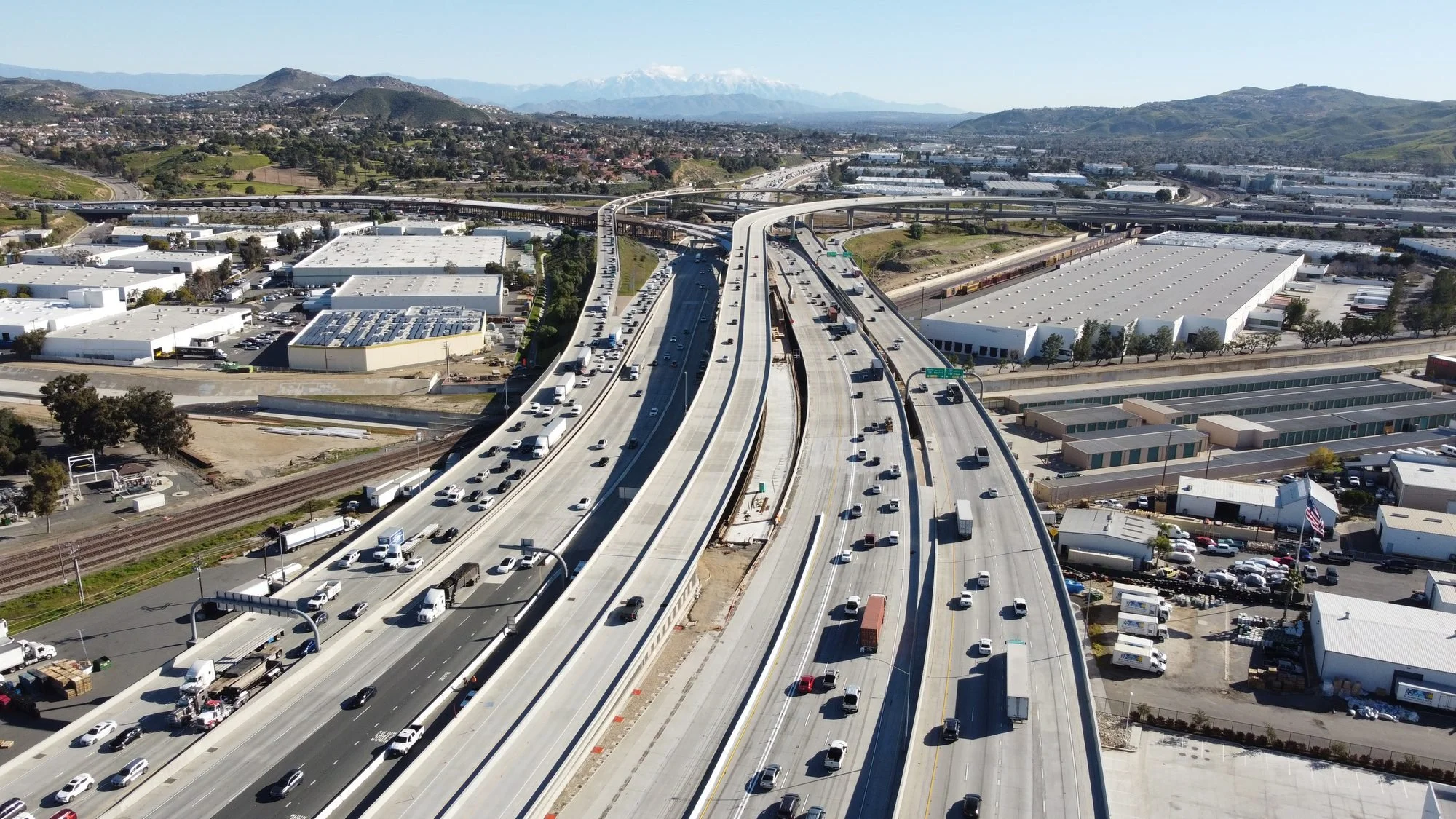 Aerial view of a busy multi-lane freeway with cars and trucks, elevated over construction areas, with industrial buildings nearby, hills, and mountains in the background.