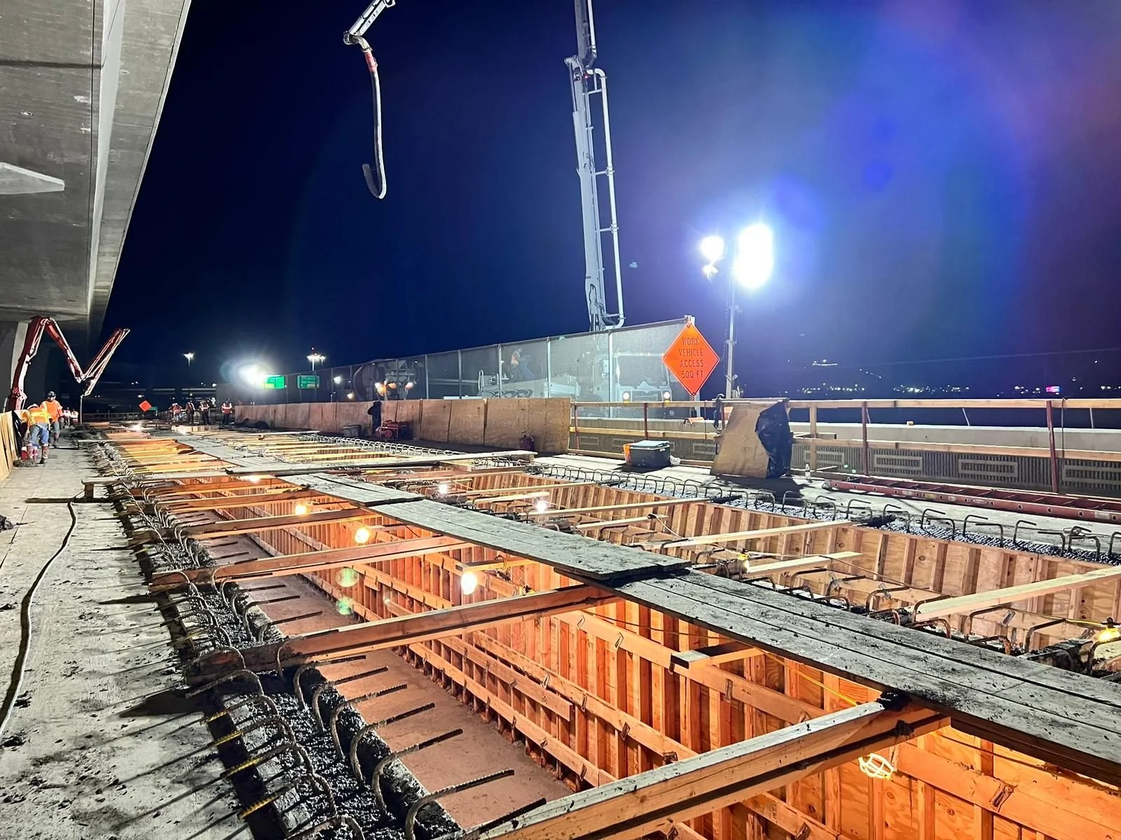 Nighttime construction site on a highway, workers installing concrete forms for a new bridge or barrier, illuminated by bright artificial lights.
