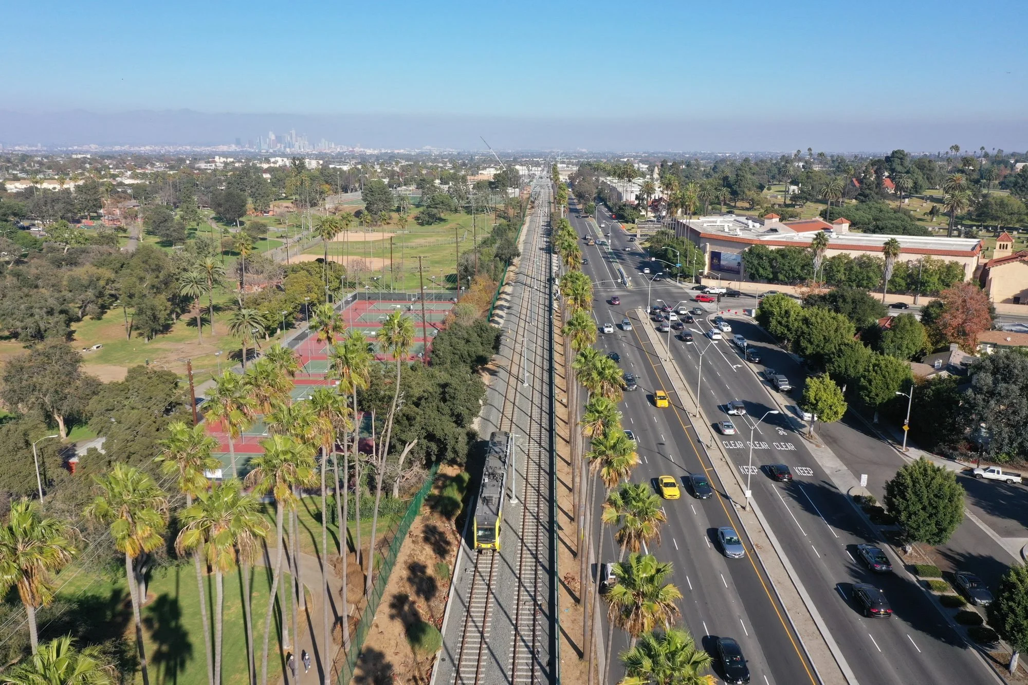 Urban landscape with a park, tennis courts, and a busy road surrounded by palm trees and green spaces, with a city skyline in the background.