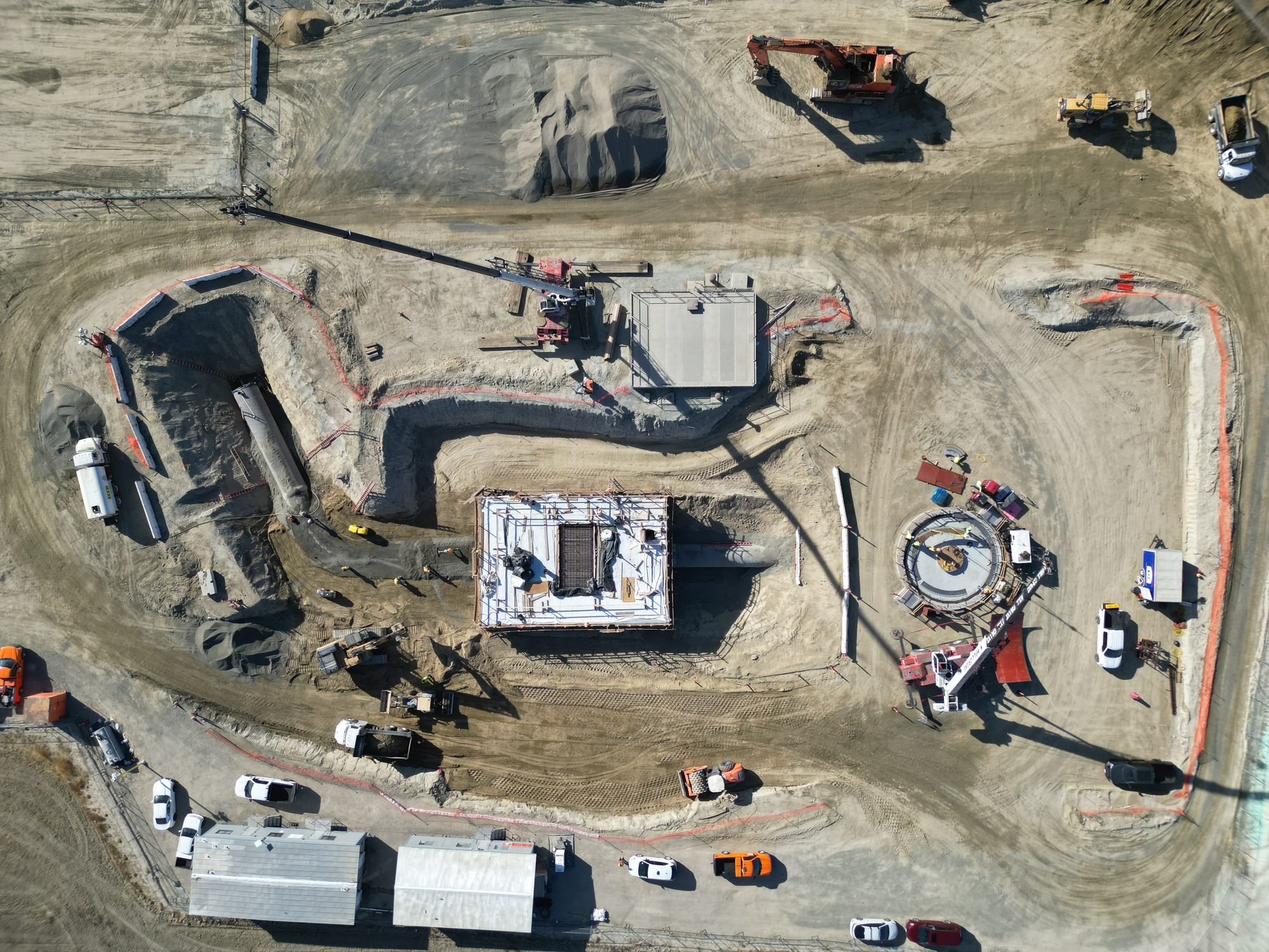 An aerial view of a construction site with heavy machinery, construction vehicles, and a partially built structure. The area has dirt roads, piles of dirt, and construction equipment.