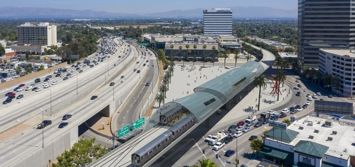 An aerial view of a modern urban area with a city train station and a train arriving at the platform, surrounded by tall buildings, a busy highway, parking lots, and trees.