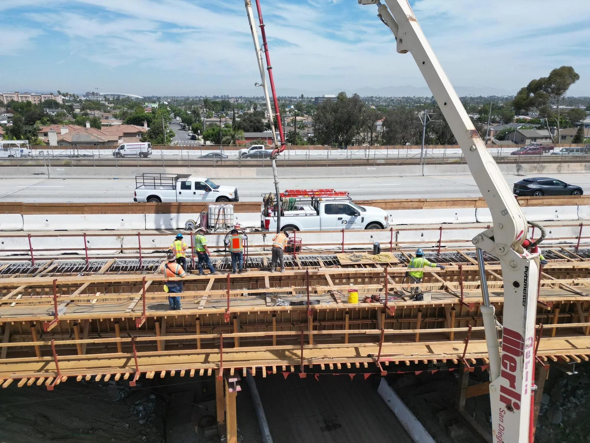Construction workers working on a bridge or overpass, with vehicles passing below, and a cityscape in the background.
