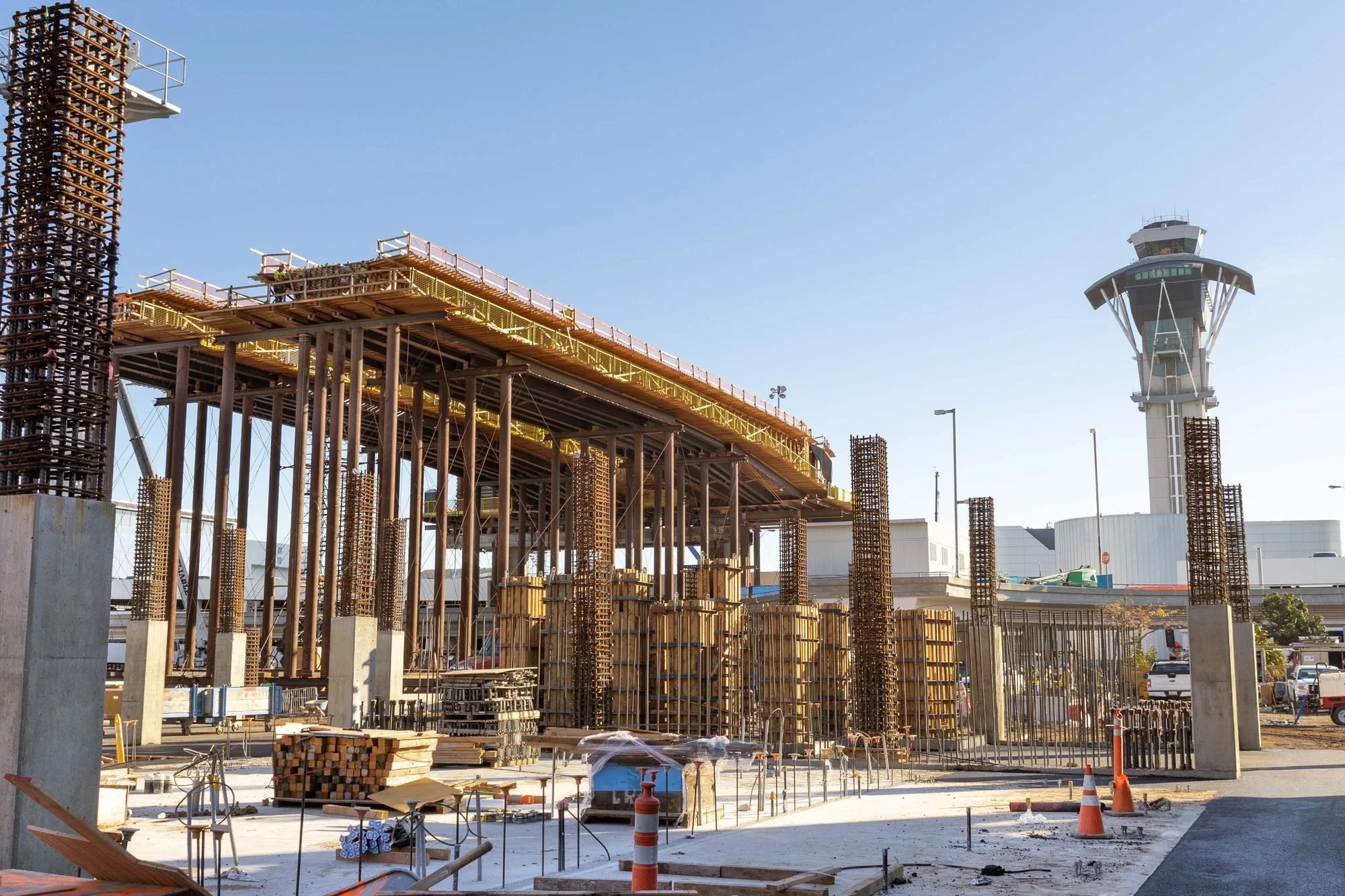 Construction site with building framework, steel rebar, and a control tower in the background under a clear blue sky.