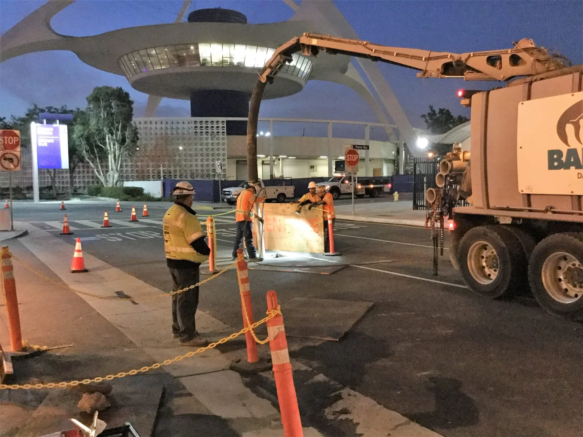 Workers in safety vests and helmets working on a construction site, digging trenches with equipment, near a modern building during dusk.