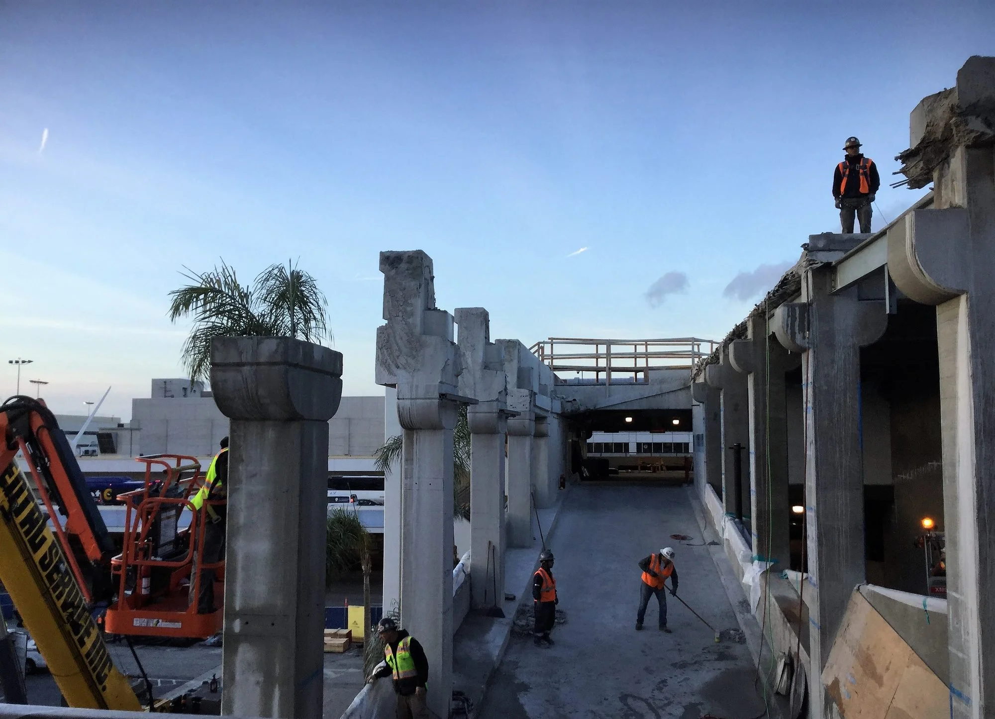 Construction workers with safety gear working on a building structure at dusk, with a clear sky and palm tree in the background.