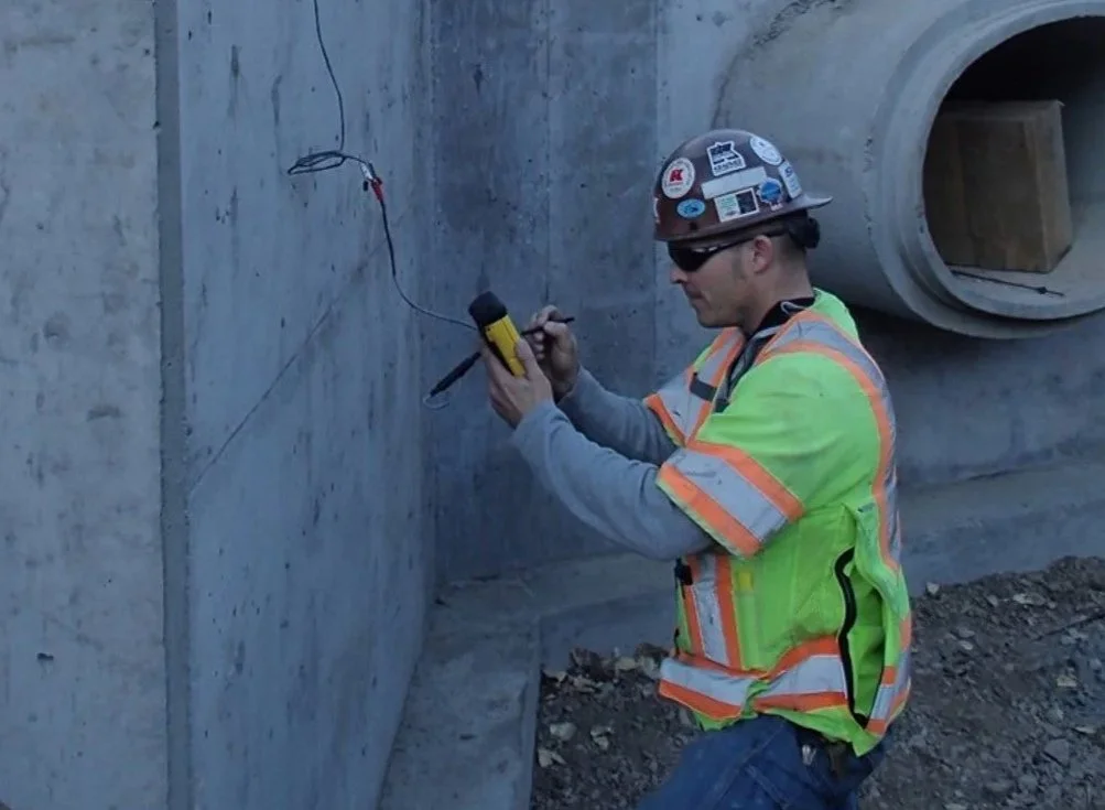 Construction worker wearing a safety helmet and vest, inspecting concrete pipe underground using a measuring device at a construction site.