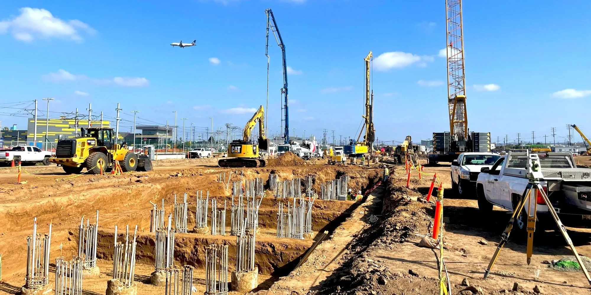 Construction site with excavators, trucks, and workers, with an airplane flying in the clear blue sky overhead.