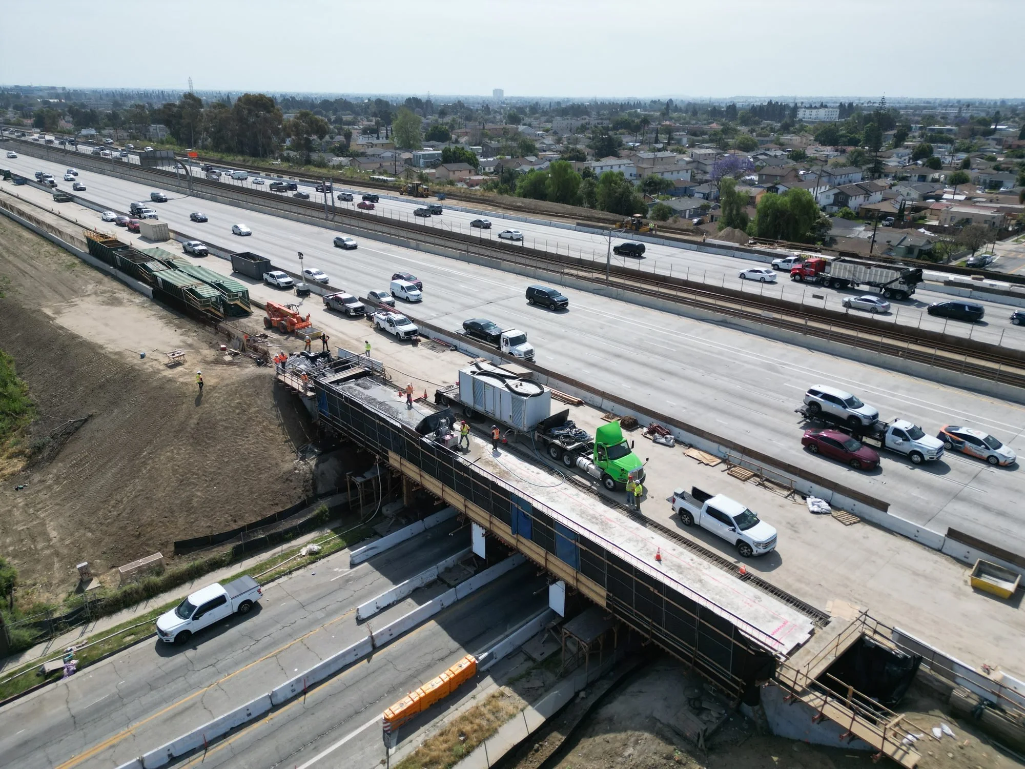 Elevated view of a highway construction site with workers and machinery, next to a residential neighborhood with houses and trees under a clear sky.