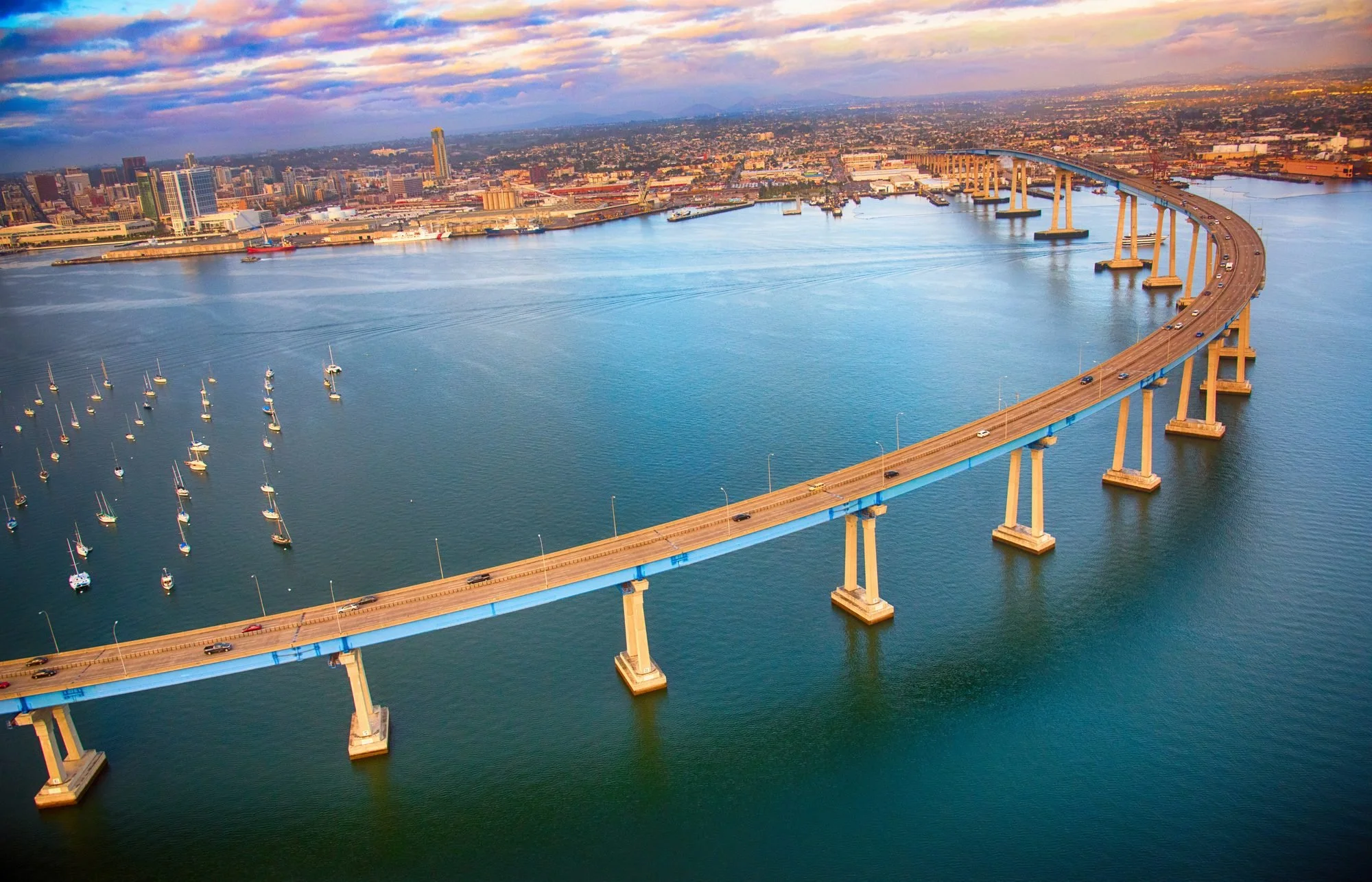 An aerial view of a large city with a bridge over a wide body of water. The city skyline includes many high-rise buildings, and the water has numerous sailboats and a few ships. The sky is partly cloudy with colorful clouds.