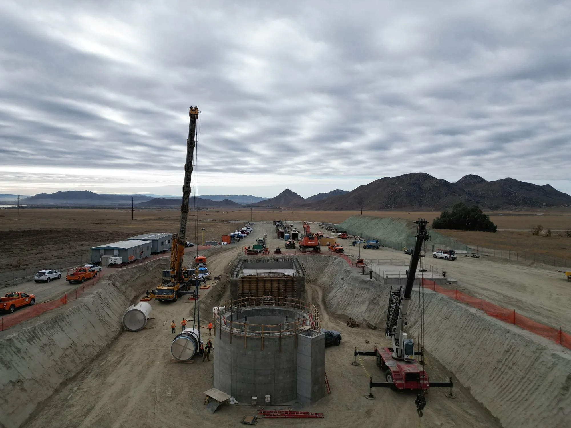 Construction site with large cylindrical concrete structures, cranes, construction vehicles, workers, and mountainous landscape in the background under cloudy skies.