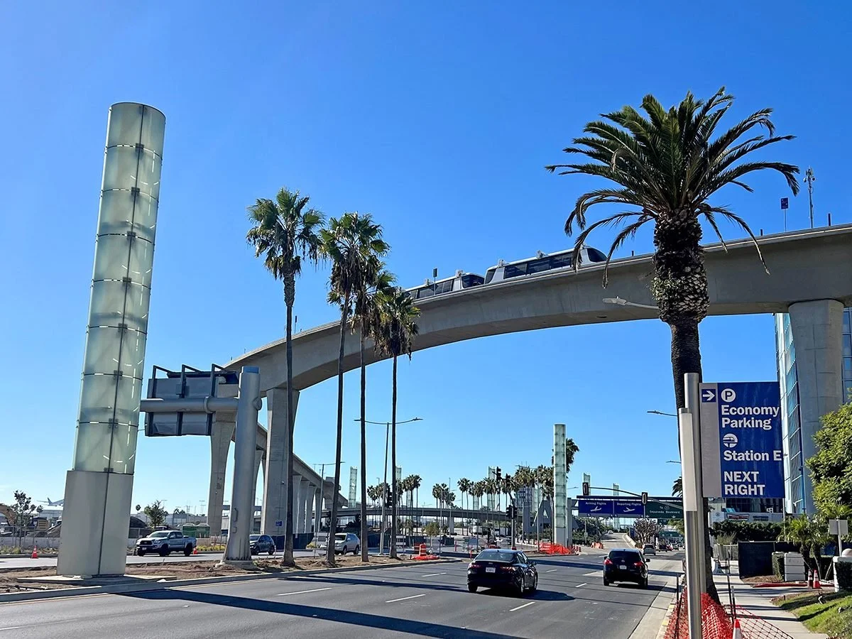 A city street with palm trees, a curved elevated train track, cars driving, and signs indicating parking and station directions in a sunny, clear sky.
