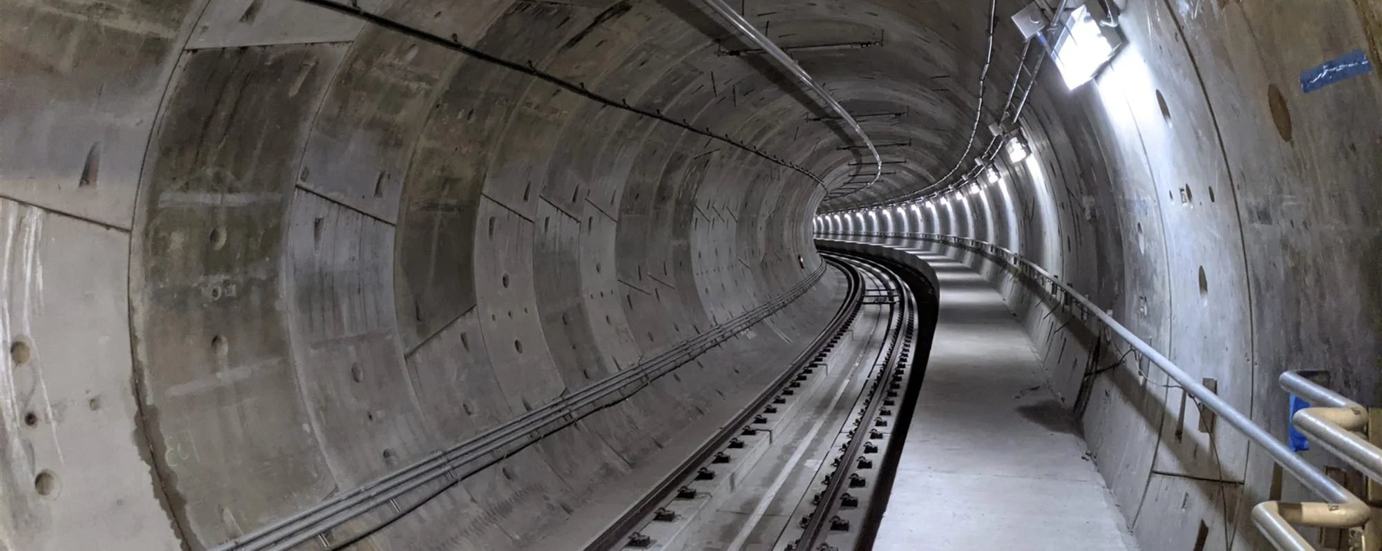 Inside a long, curved underground train tunnel with tracks and overhead lighting.