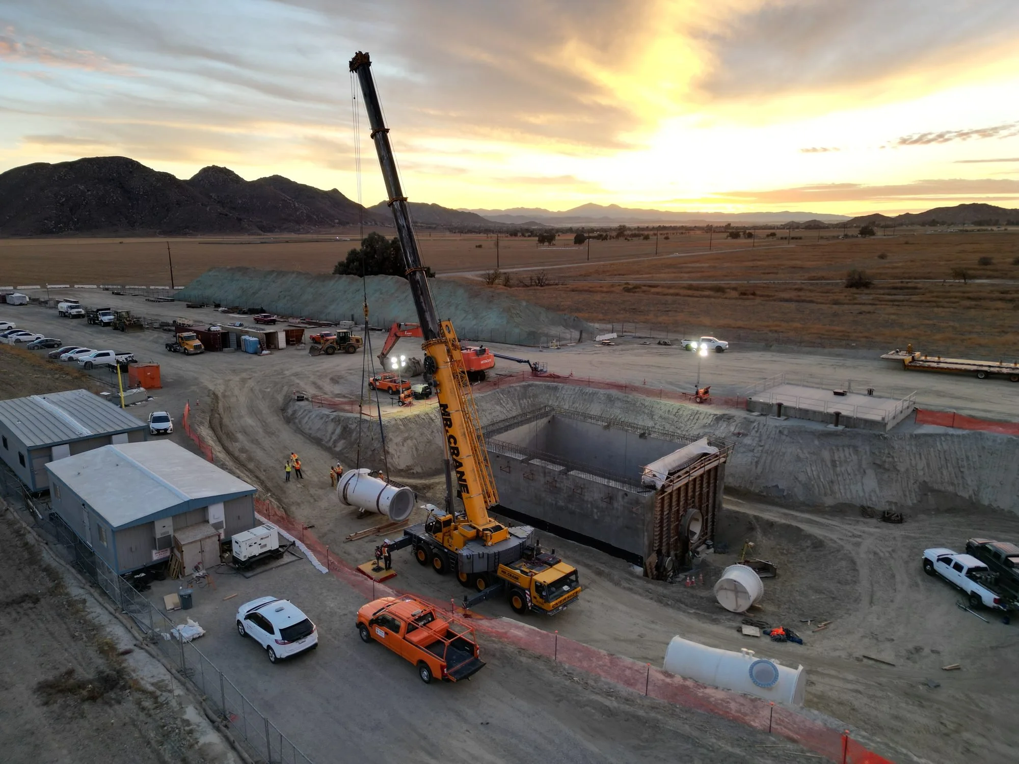 Construction site in a desert area with construction vehicles, a crane, and partially built structures during sunset.