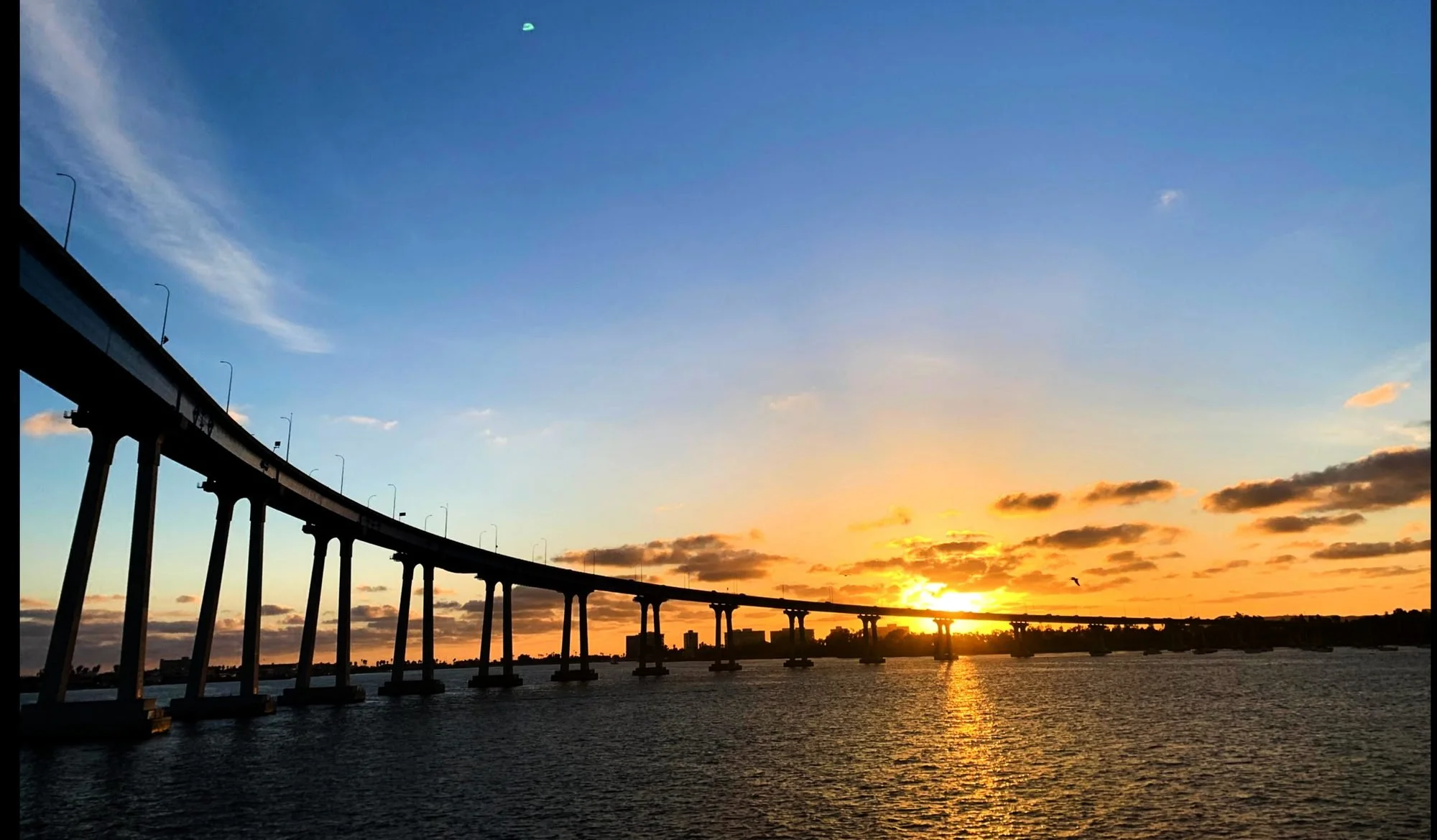 A bridge over water during sunset with a colorful sky, clouds, and a bird flying.