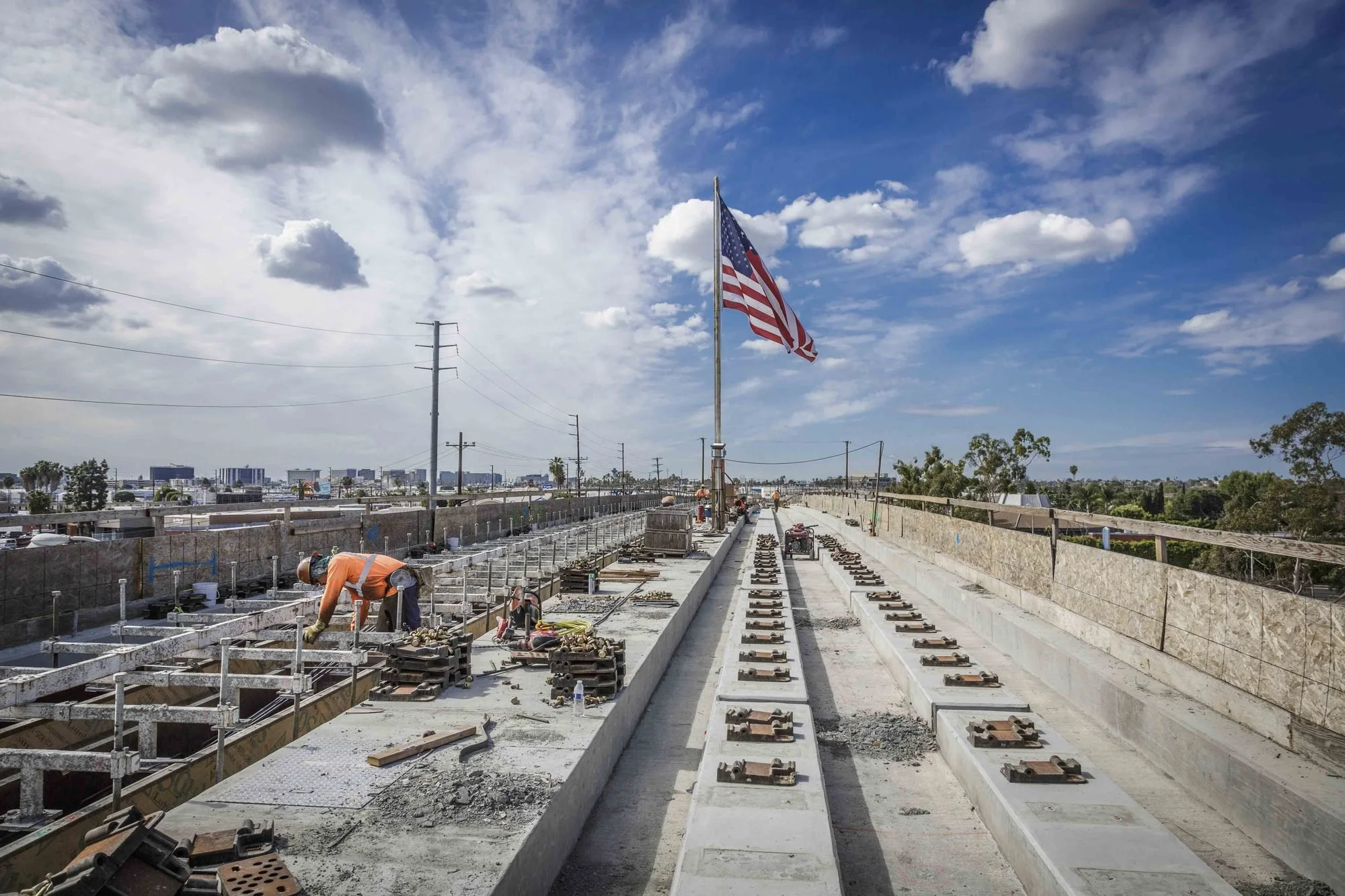 Construction workers laying down track for a new train or tram line on an overpass with a city skyline in the background. An American flag is flying on a pole in the center of the image.