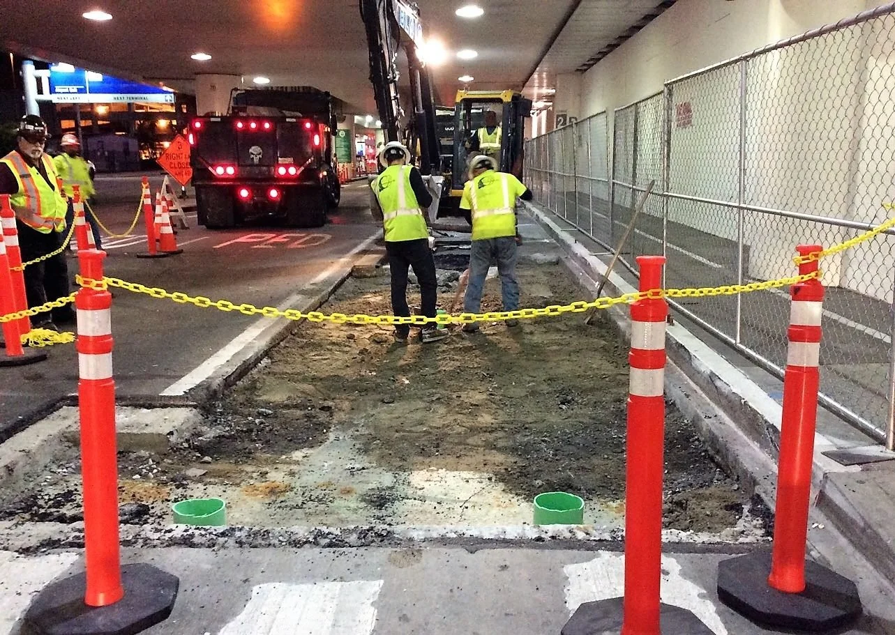 Construction workers in safety vests and helmets working on a sidewalk under an overpass, with orange cones and yellow caution tape preventing access.