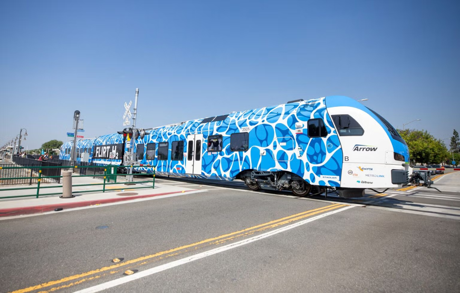 A modern blue and white commuter train stopped at a station, with artwork resembling water ripples and multiple bulbs on the front. The station platform has benches and a railroad crossing signal nearby, under a clear blue sky.