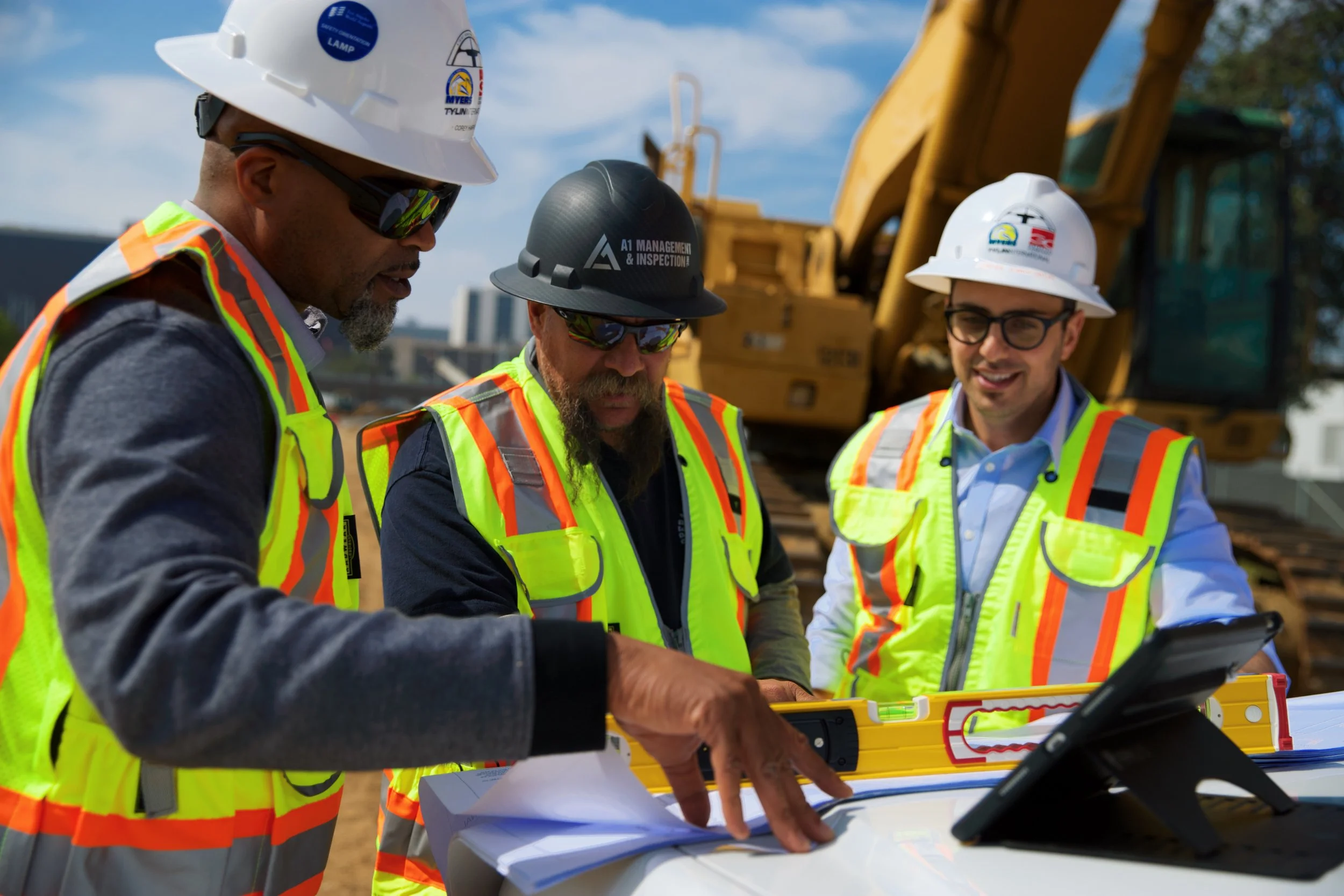 Three construction workers wearing safety helmets and high-visibility vests review plans and use surveying equipment at a construction site with heavy machinery in the background.