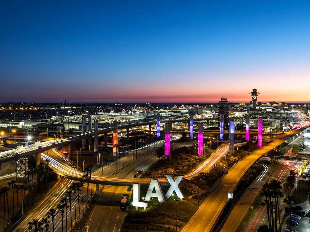 Nighttime aerial view of Los Angeles International Airport (LAX) showing illuminated roads, terminal buildings, control towers, and colorful lighting on airport support pillars with a sunset sky in the background.