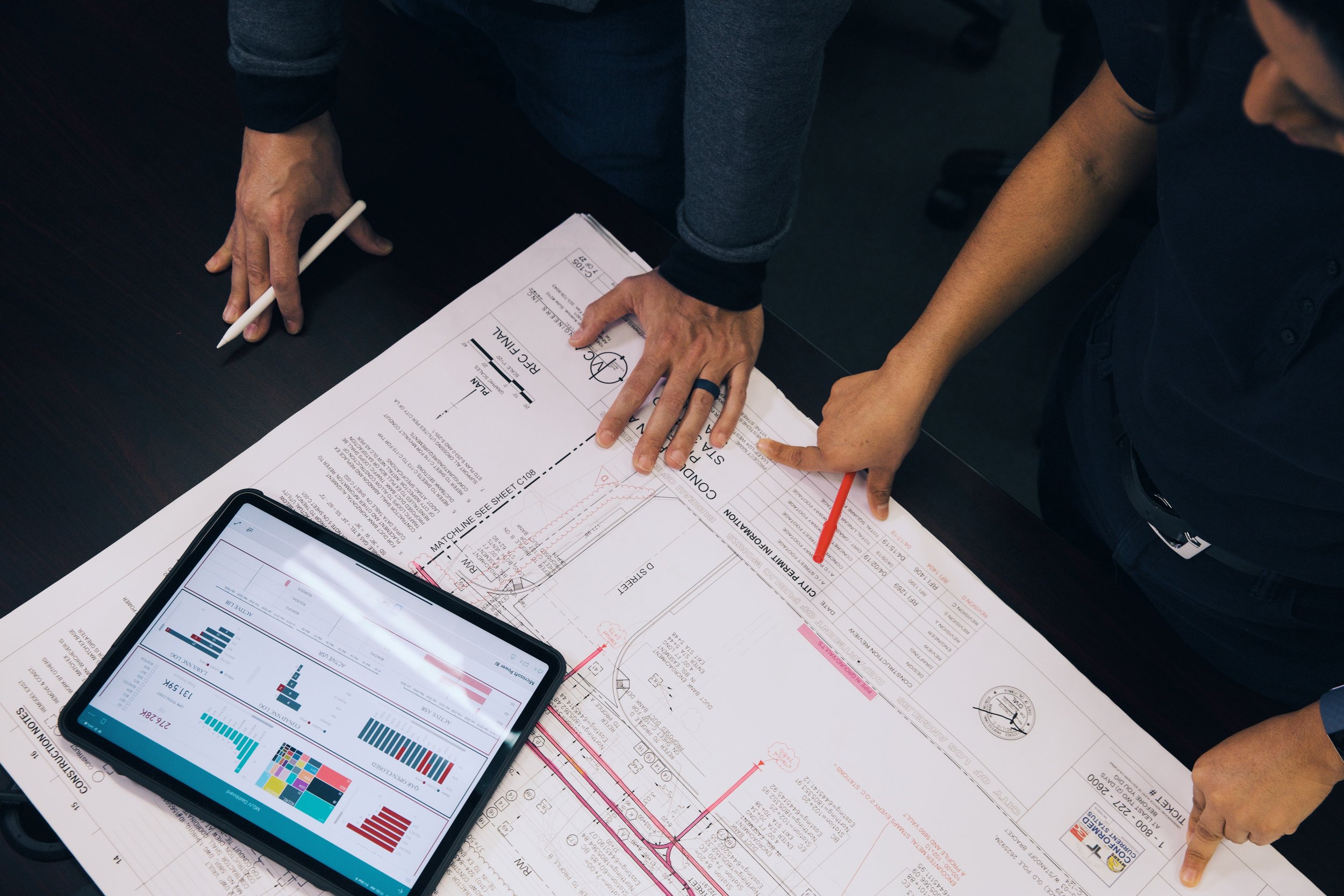 Two people examining construction or engineering plans on a table with a tablet showing charts and graphs, and a pen in one person's hand.