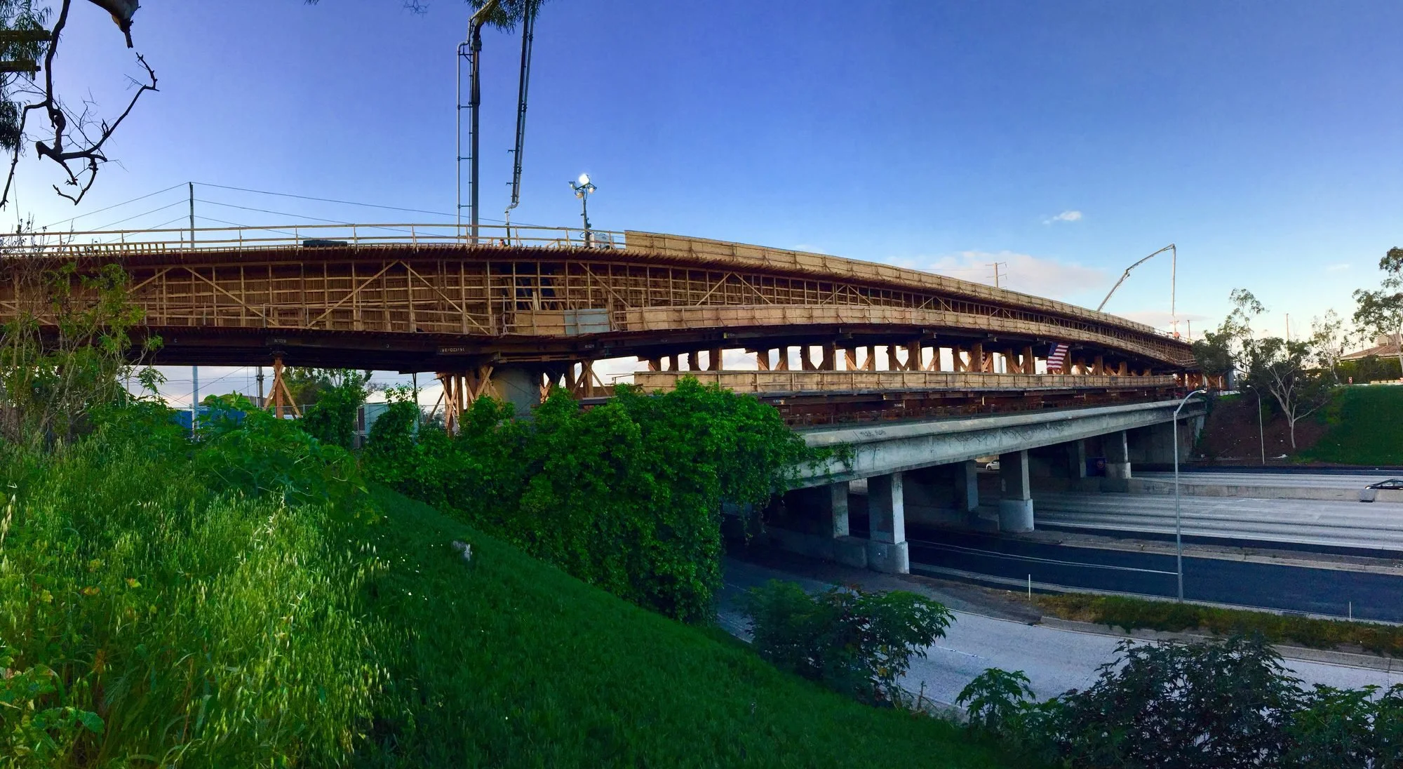 A bridge under construction over a highway with green shrubbery and trees in the foreground, and a blue sky in the background.