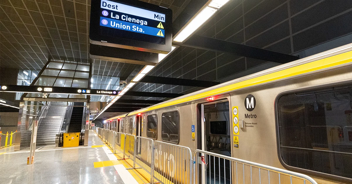 Underground metro station platform with train present. Digital sign overhead shows destination as La Cienega and Union Station, with warning signs. Escalator and stairs are visible.