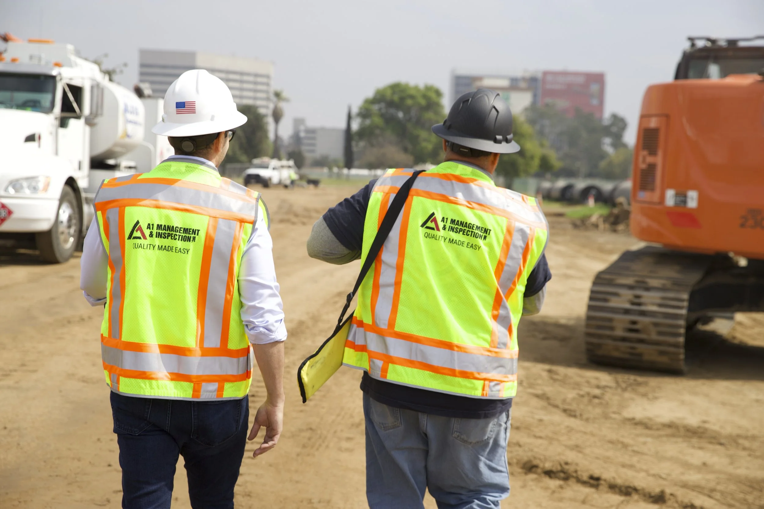 Two construction workers wearing yellow safety vests and hard hats walking on a construction site, with large machinery and a cityscape in the background.