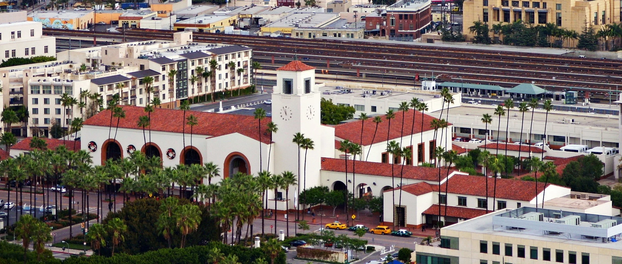 Aerial view of a cityscape with a prominent white building featuring a clock tower, surrounded by palm trees, modern buildings, and train tracks in the background.