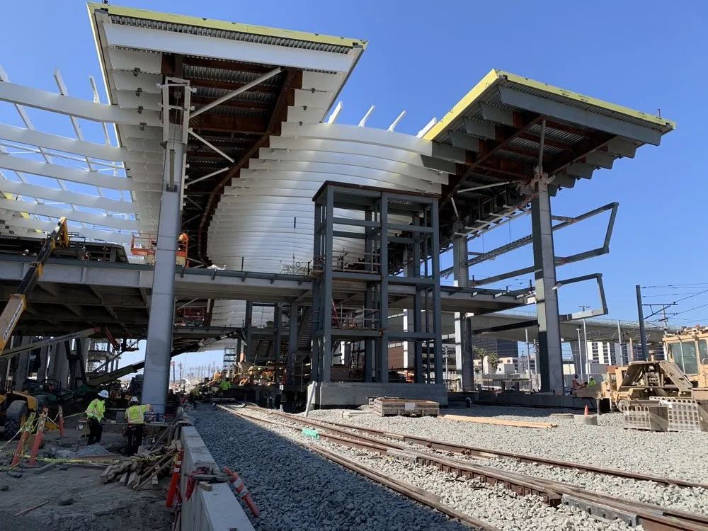 Construction site of a modern building with steel framework and overhanging roof sections, near train tracks, with workers and construction equipment present.