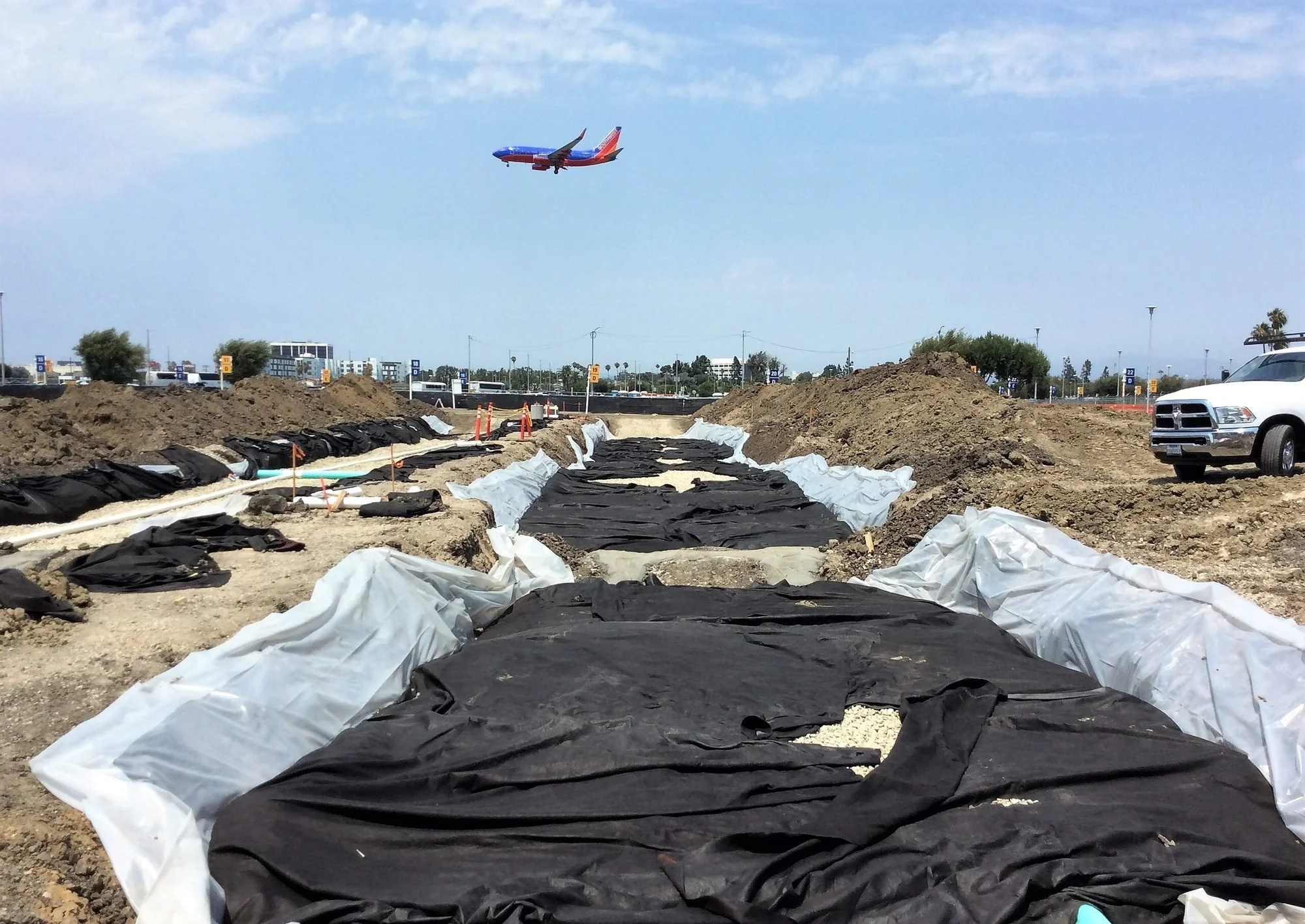 Construction site with black and white plastic sheets covering the ground, dirt mounds, and an airplane flying in a partly cloudy sky.