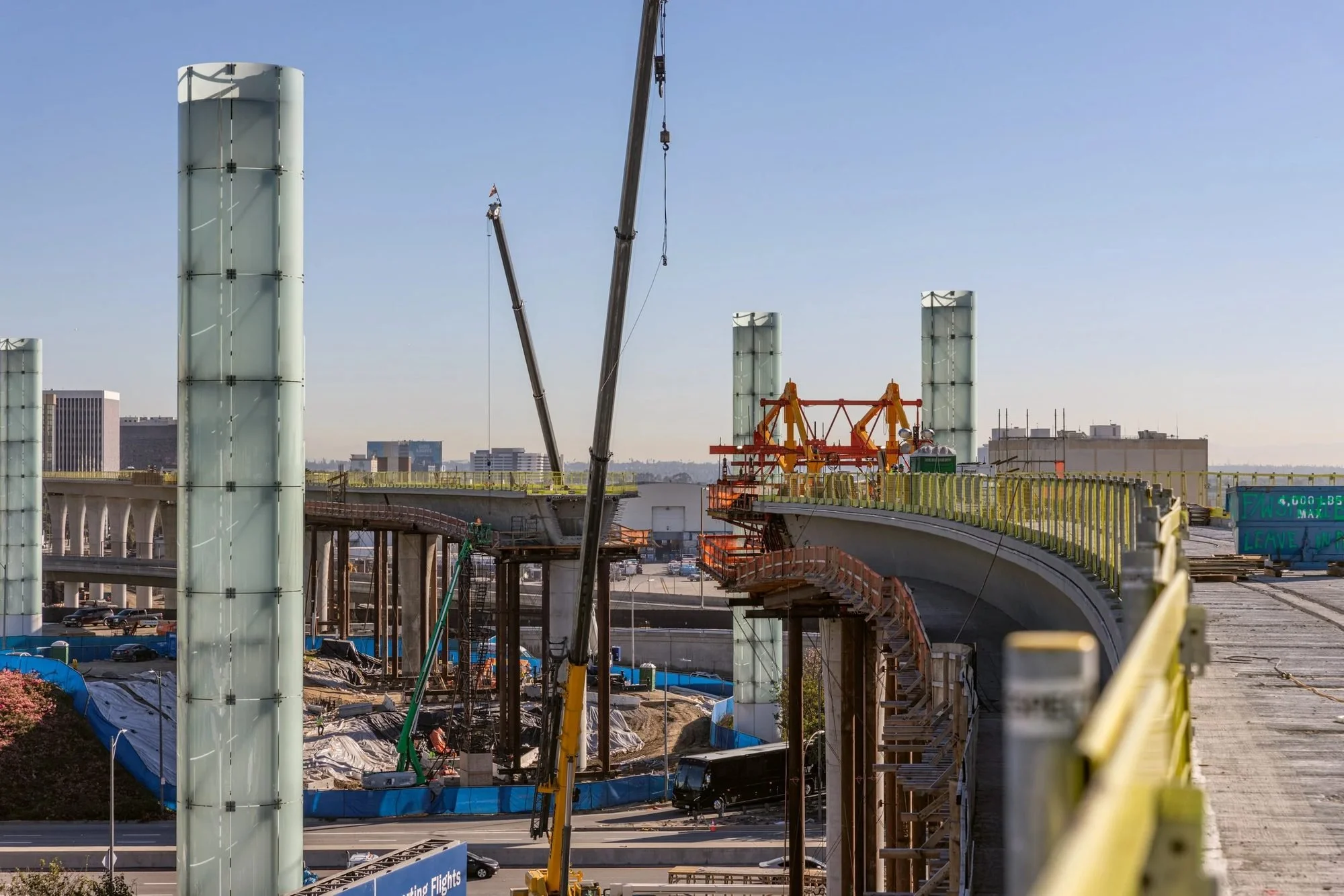 Construction of a new highway or overpass with large glass panels being installed and cranes in use, surrounded by safety barriers and construction equipment.