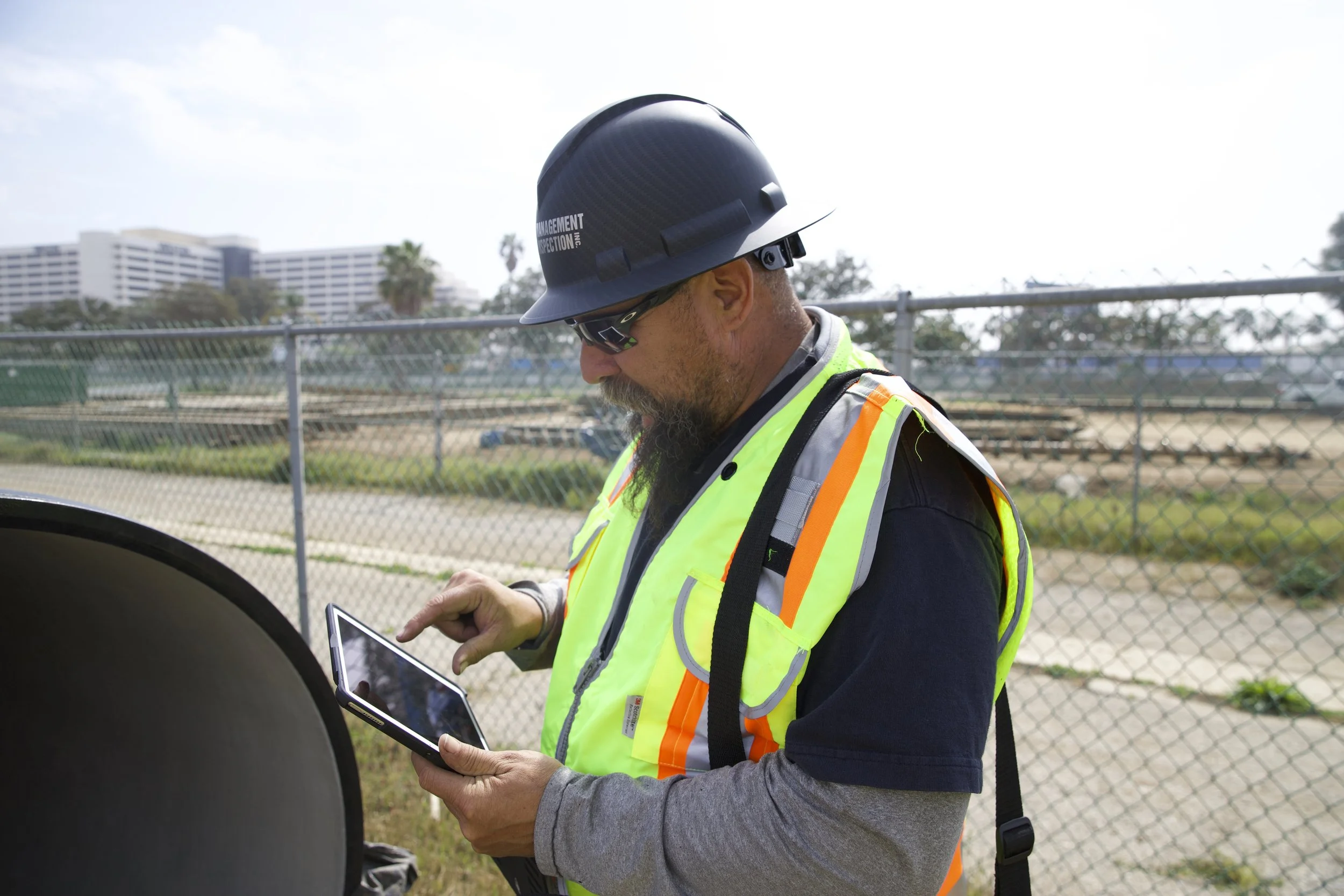 A man wearing a black safety helmet and sunglasses, dressed in a gray long sleeve shirt and a reflective safety vest, holding and looking at a tablet device outdoors near a chain-link fence and city buildings in the background.