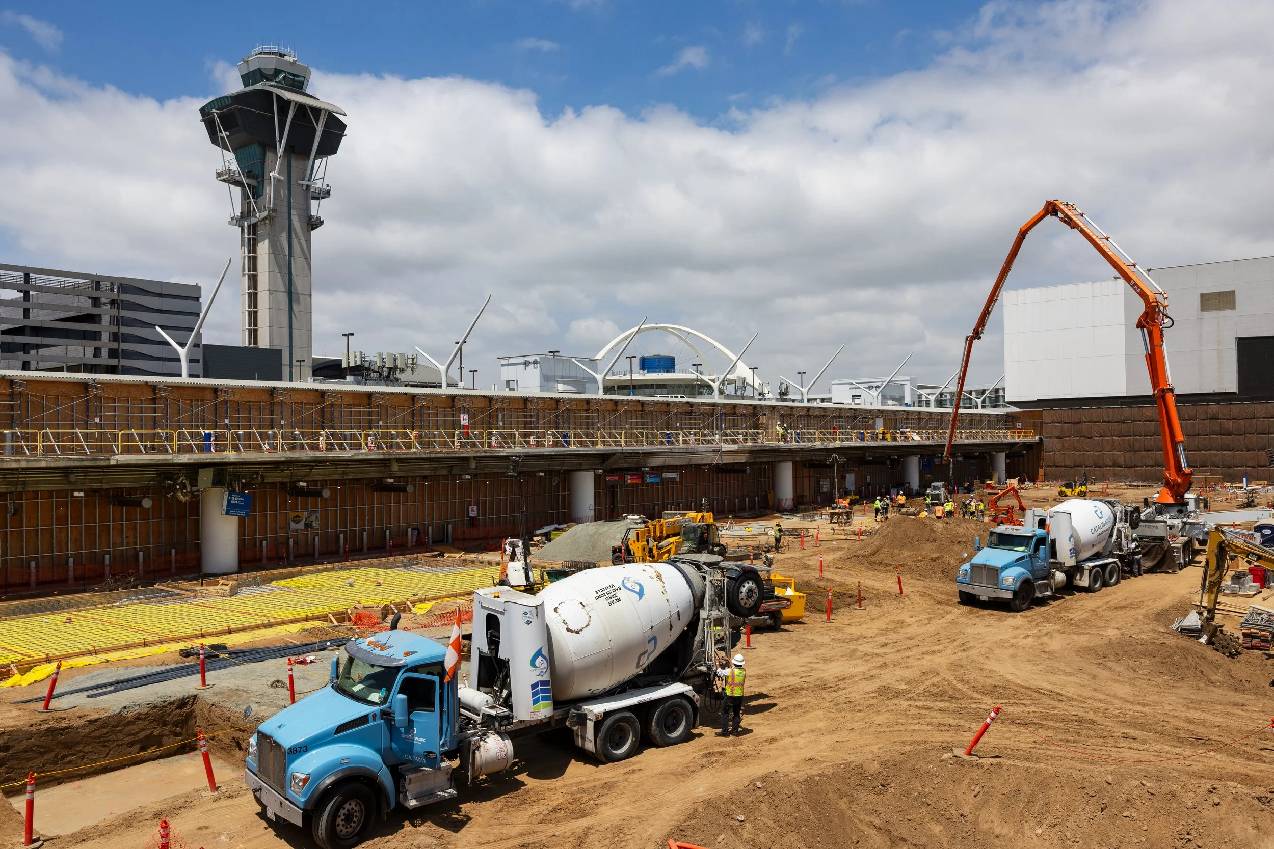 Construction site at an airport with a control tower in the background. Several construction vehicles, workers, and equipment are visible, with scaffolding and a concrete pour area in the foreground.