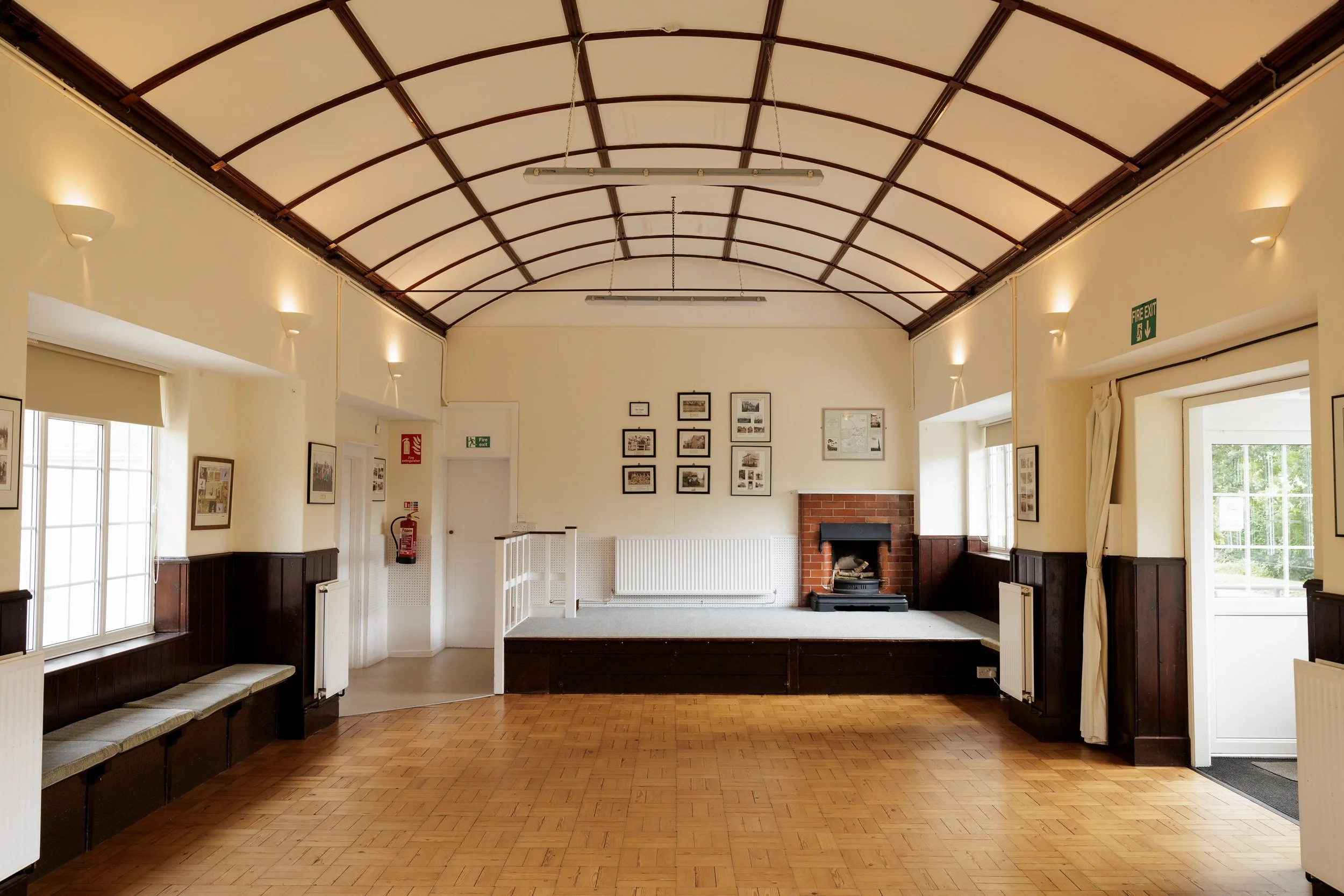 A spacious room with wooden flooring, windows on both sides, a small stage with a brick fireplace, and framed pictures on the wall.