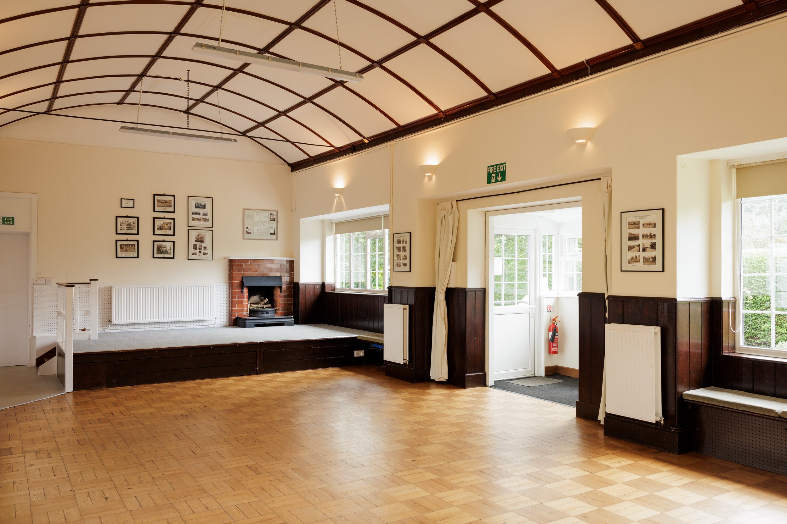 A spacious room with wooden flooring, white walls, and a curved ceiling with wooden beams. There are several framed pictures on the walls, a brick fireplace, and large windows letting in natural light. A fire exit door and a fire extinguisher are als