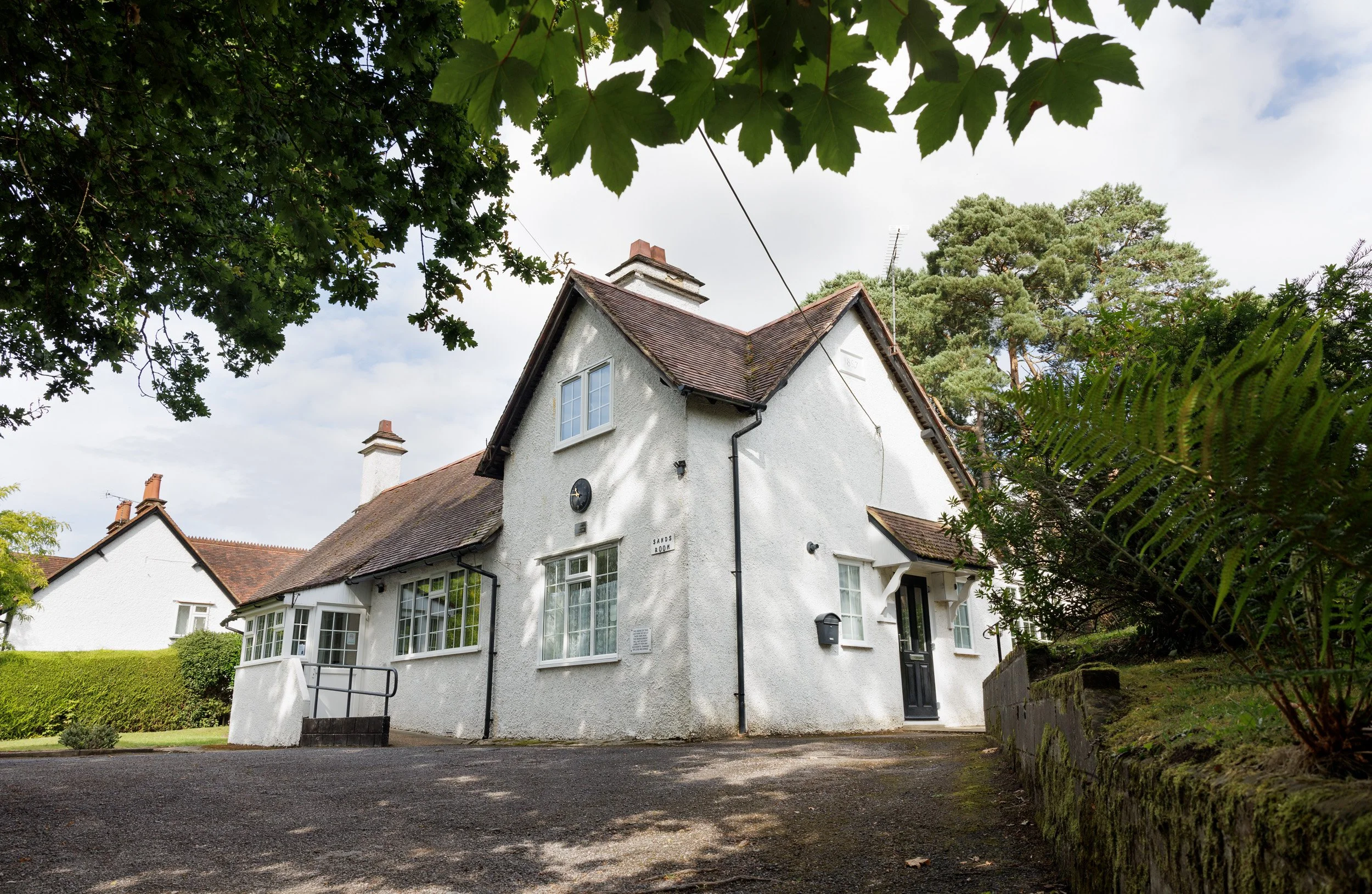 Exterior of The Sands Room village hall, multiple windows, a sloped roof, and chimneys, surrounded by greenery and trees, under a partly cloudy sky.