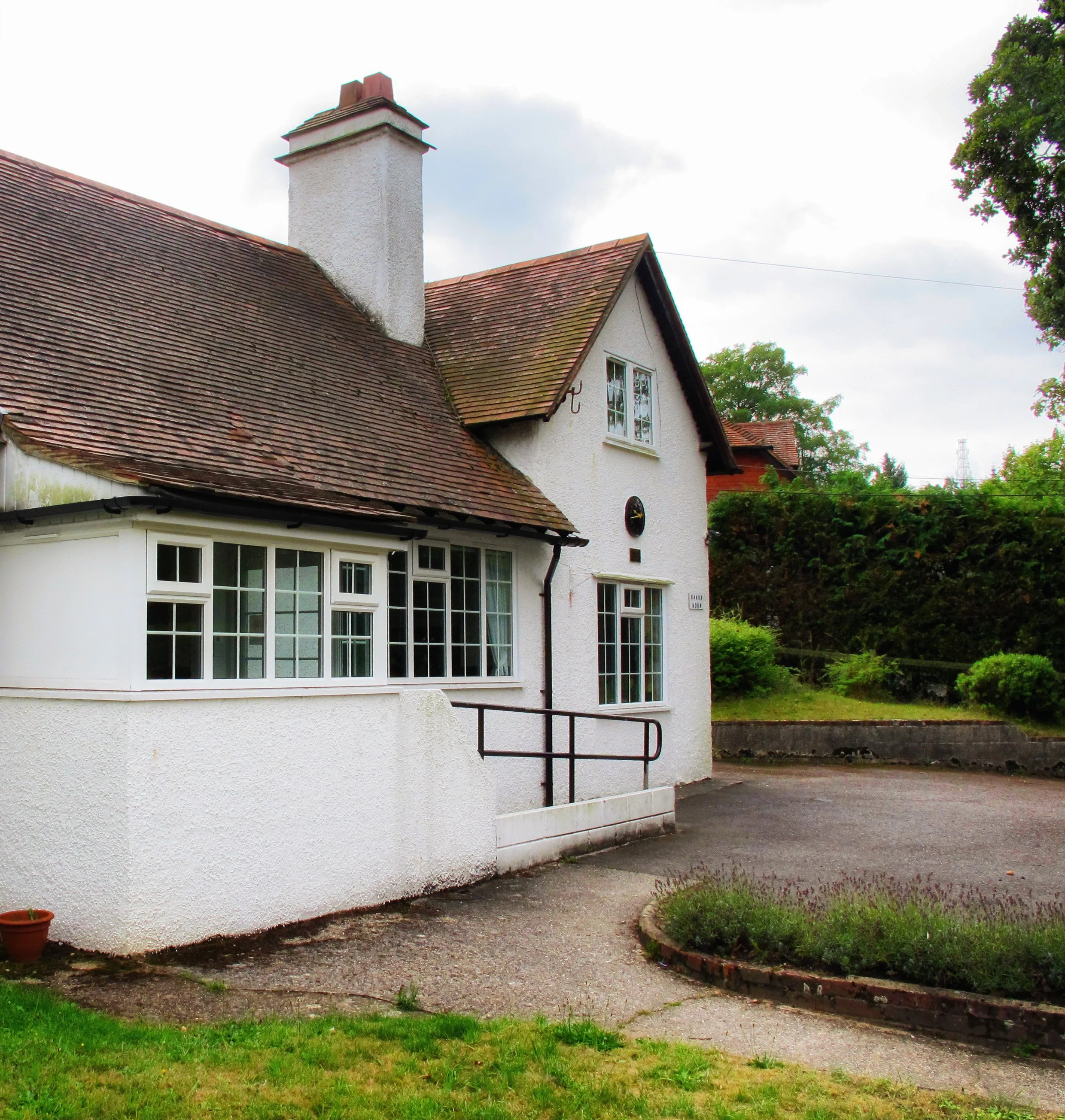 The exterior of the Sands Room village hall, a white building with large windows and a sloped roof, a chimney, a small tree in a circular garden bed, with green landscaped surroundings and a cloudy sky.