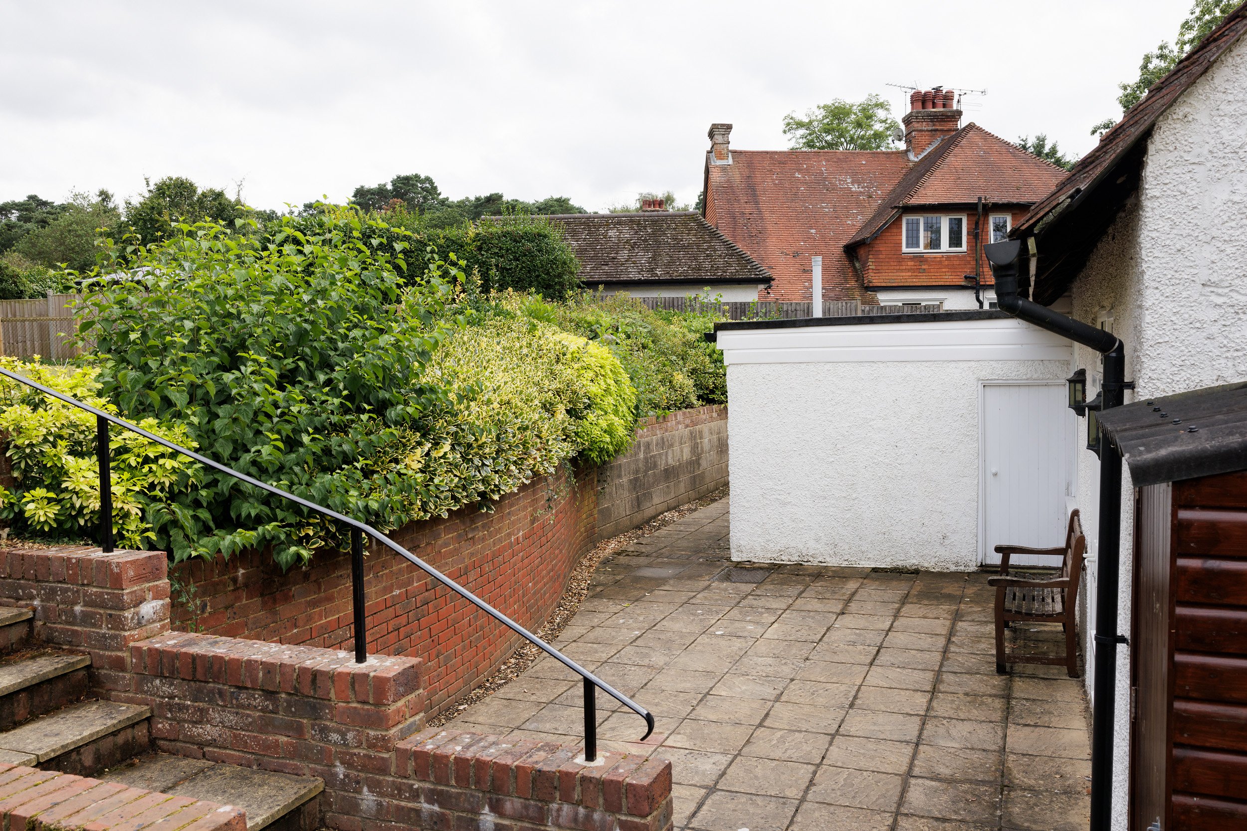 A backyard patio area with brick steps and a black metal railing, green bushes lining a brick wall, a small white building with a door, two wooden chairs, and neighboring houses with red roofs and chimneys in the background.