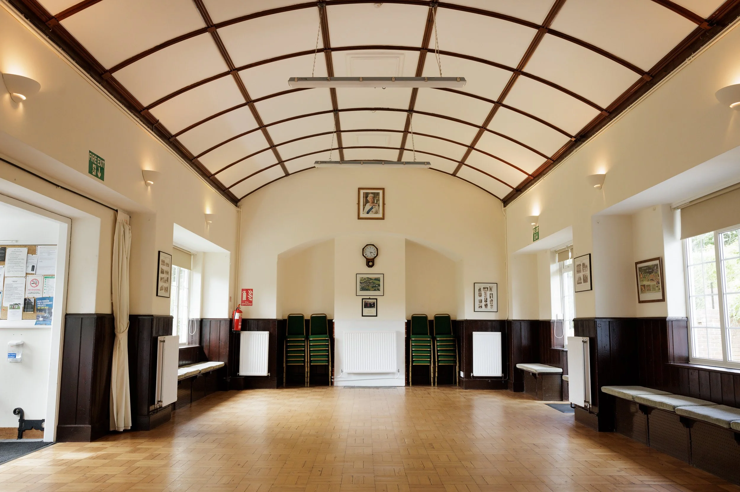 The Sands Room interior with a high arched ceiling, wood paneling walls, windows on both sides, framed pictures, a clock, stacked chairs at the back, and radiators along the walls.