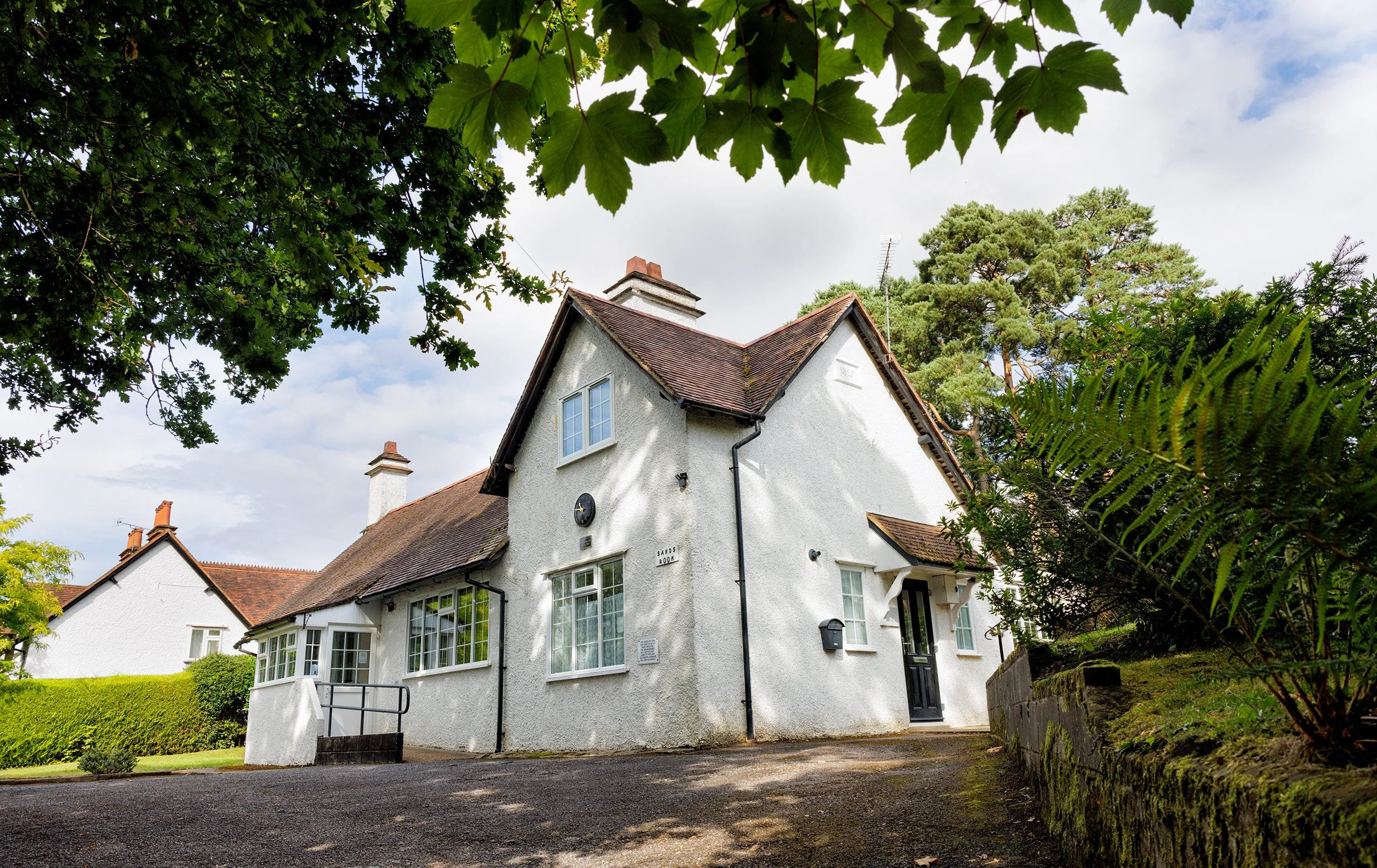The front of The Sands Room village hall with a red-tiled roof and a chimney, surrounded by green trees, with a driveway leading to the front door.