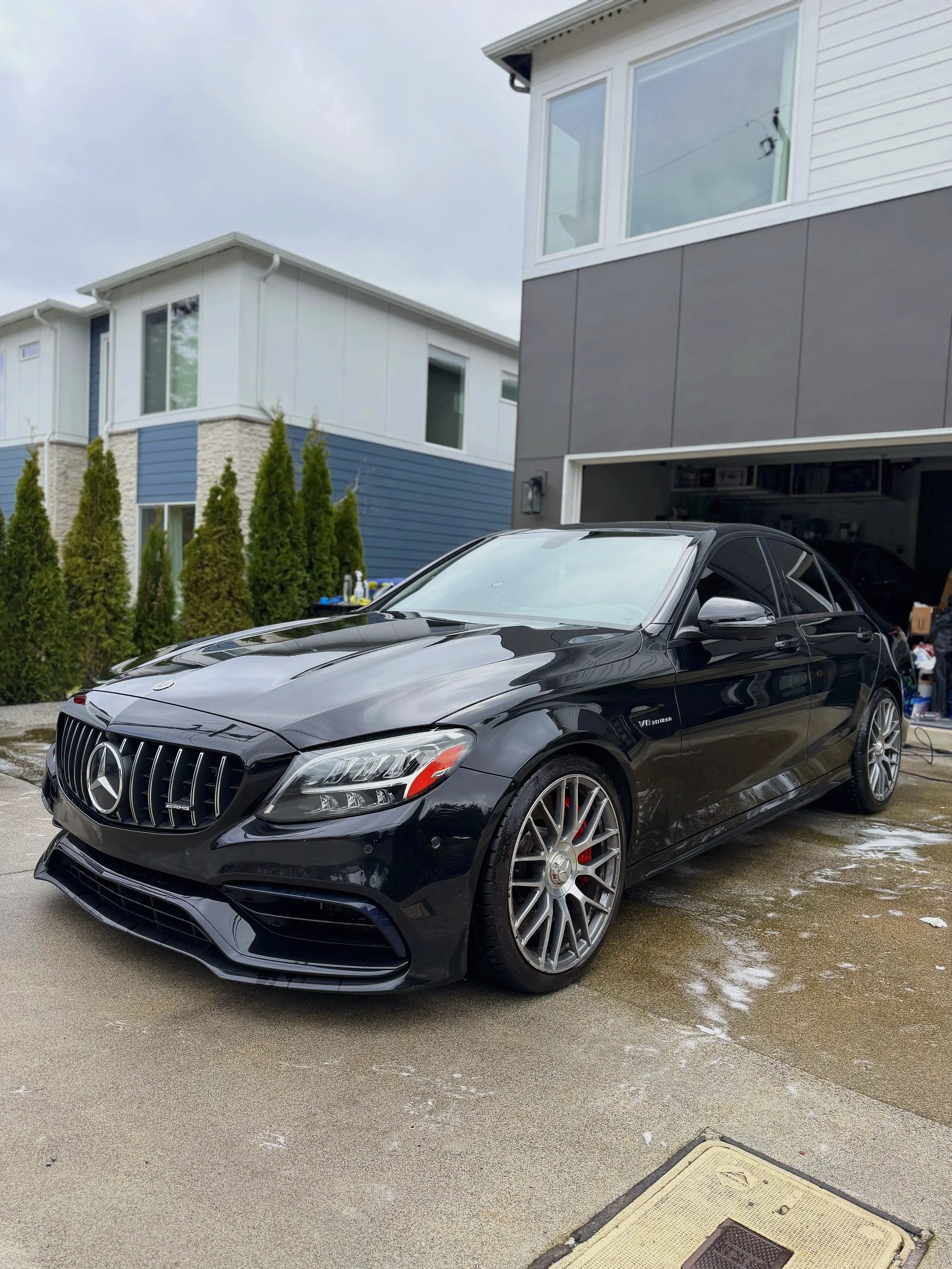 A black Mercedes-Benz AMG sedan parked in a driveway near modern houses with a cloudy sky overhead.