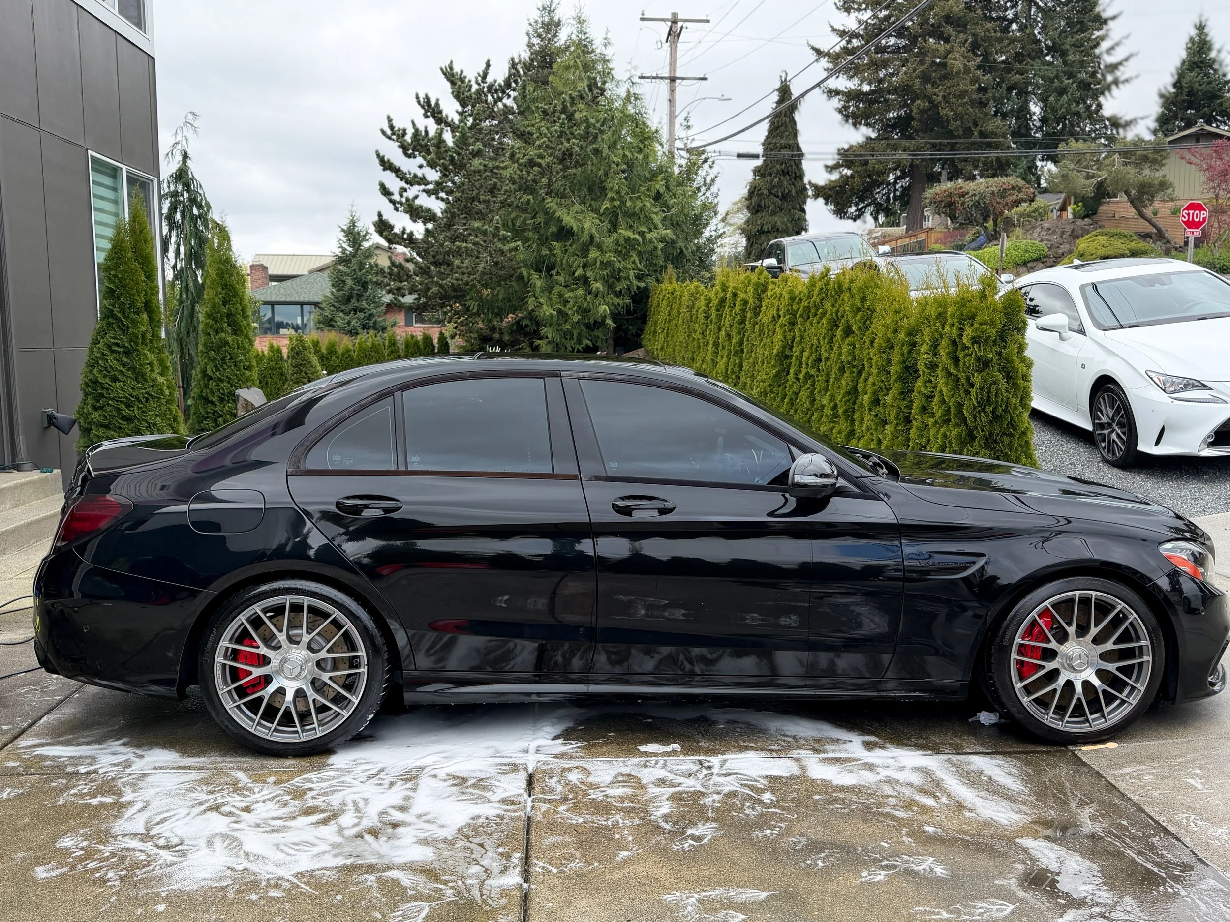 Black Mercedes-Benz sedan parked outside on a driveway with soap suds around its wheels, surrounded by green trees and other cars in the background.