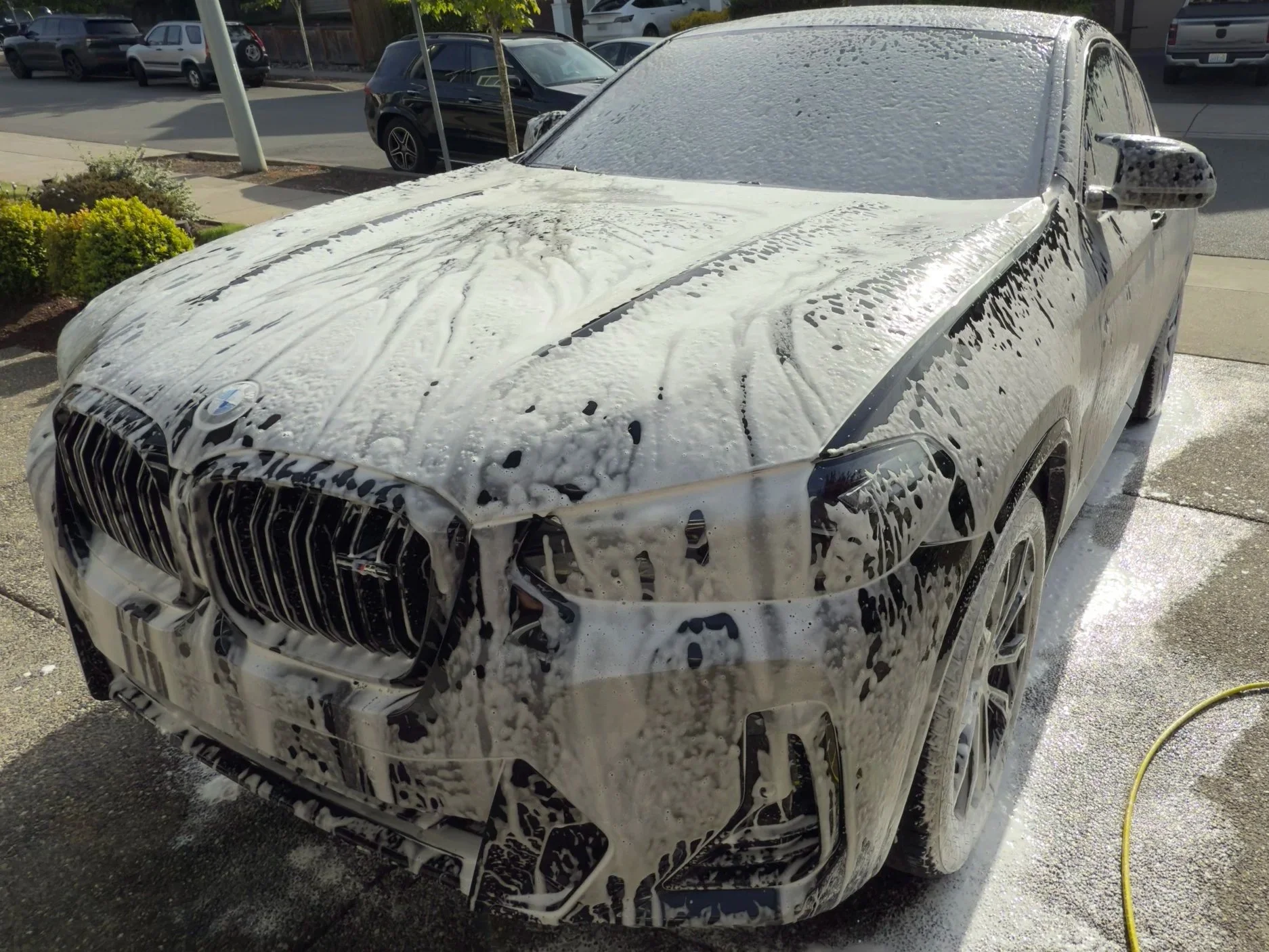 A black luxury SUV, likely a BMW, being washed with foam at a car wash. The foam covers the body, hood, windshield, and front grille, with soap suds visibly dripping down the sides.