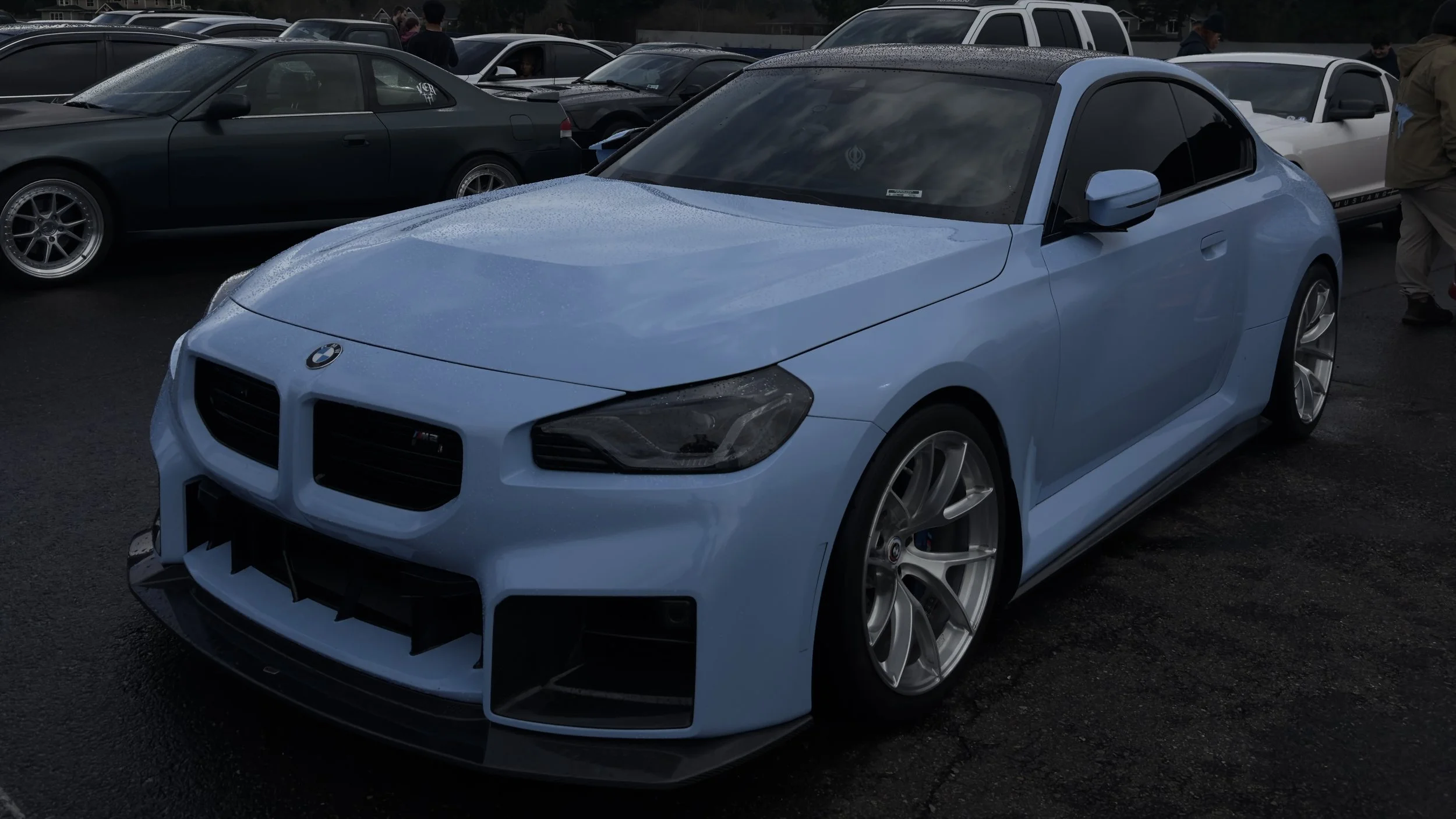 A light blue BMW M2 sports car parked among other vehicles at a car meet, with wet pavement and people in the background.