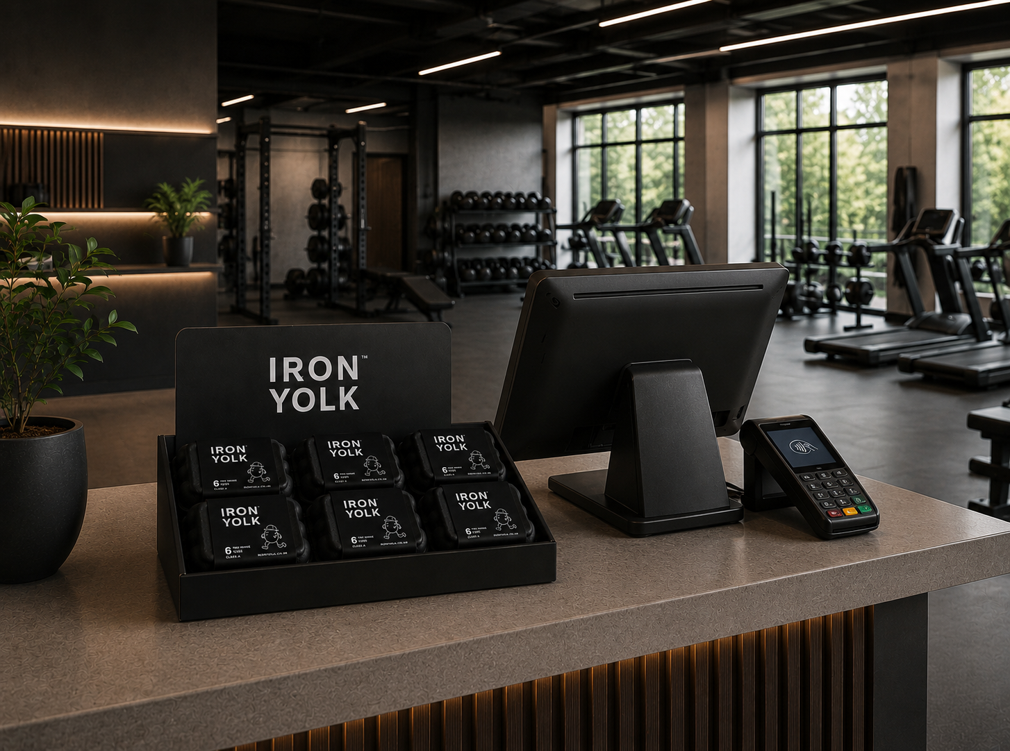 Reception desk in a modern gym with black weights and cardio equipment in the background. On the desk, there are portable food packs labeled 'Iron Yolk,' a computer monitor, a card payment terminal, and a potted plant.